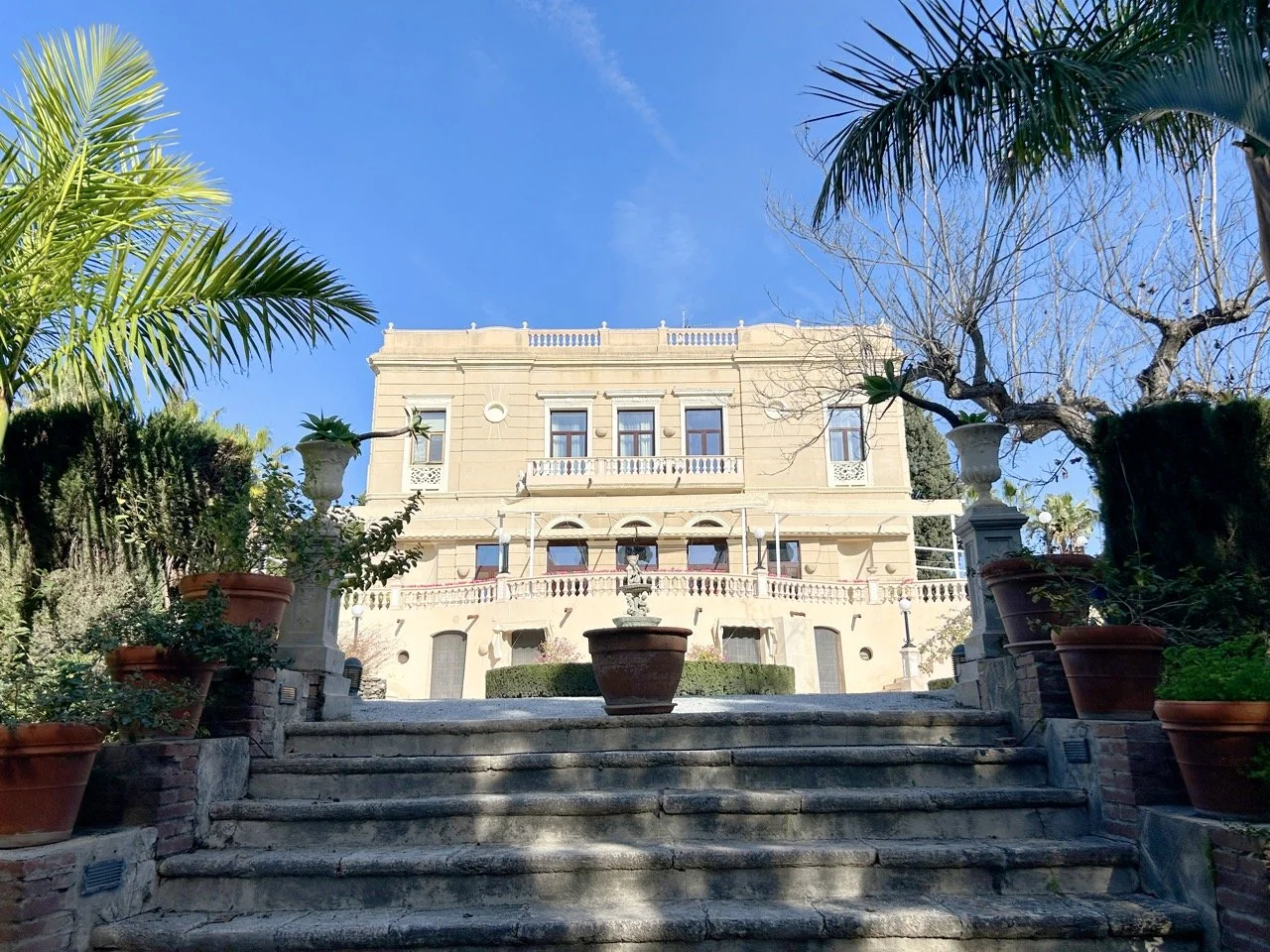 A grand beige mansion with classical architecture, surrounded by lush greenery and potted plants, with steps leading up to the entrance, under a bright blue sky.