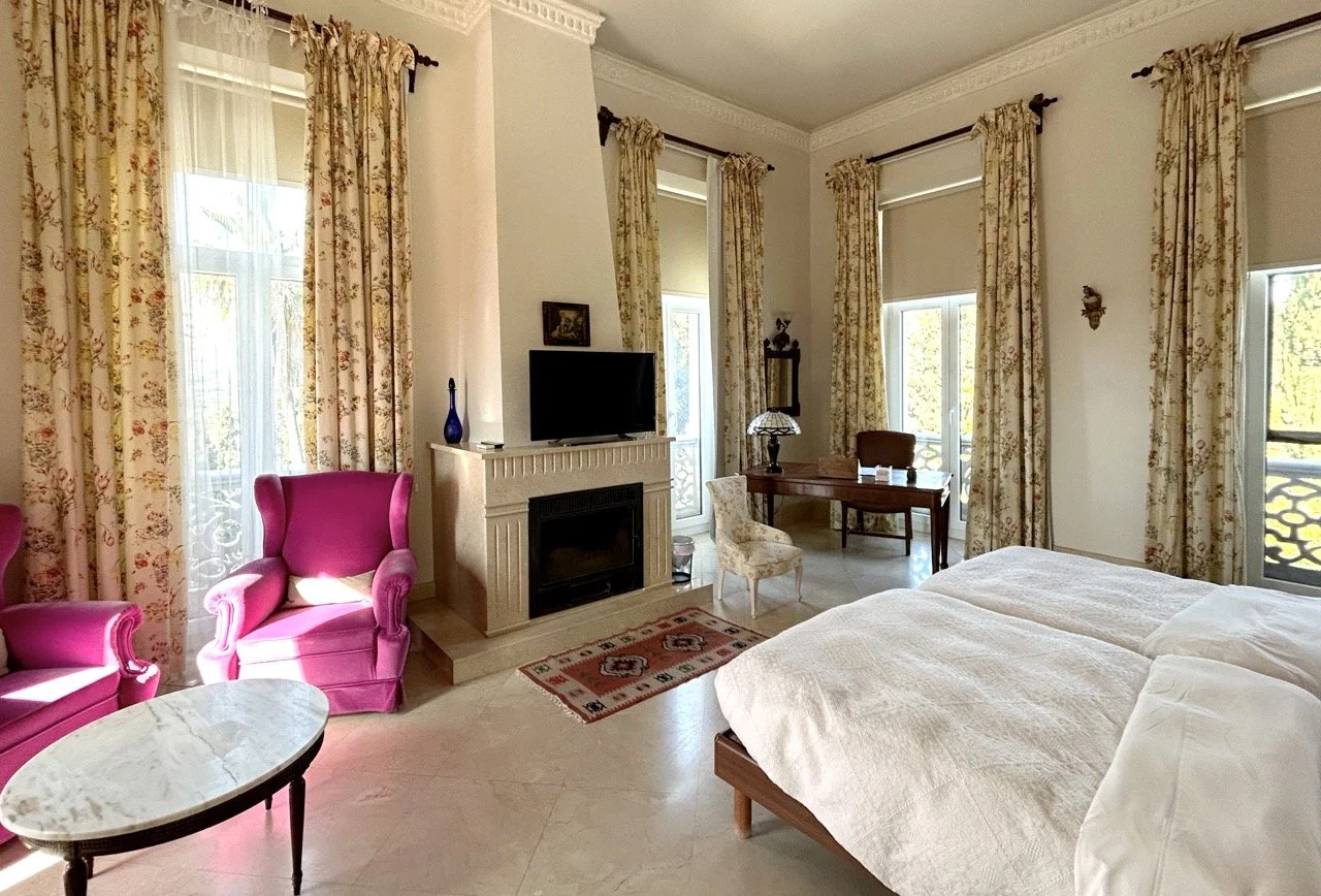 A bright hotel room with white walls and tall windows covered with floral curtains. There are two beds with white linens, a pink armchair, a small marble-topped table, a desk with a chair, a fireplace with a TV on top, and decorative items.