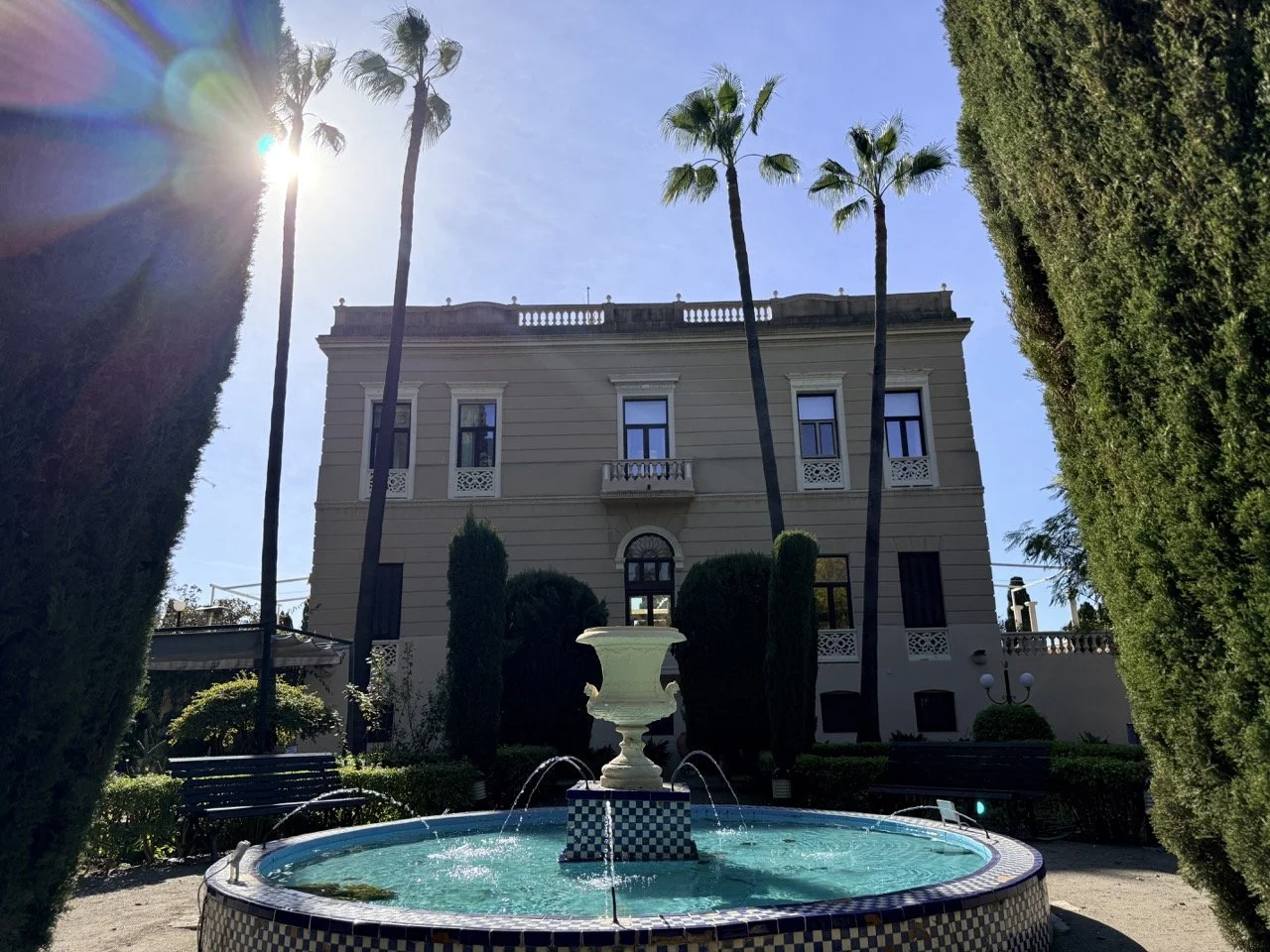 Sunlit historic building with a fountain in the foreground, surrounded by tall palm trees and lush greenery.