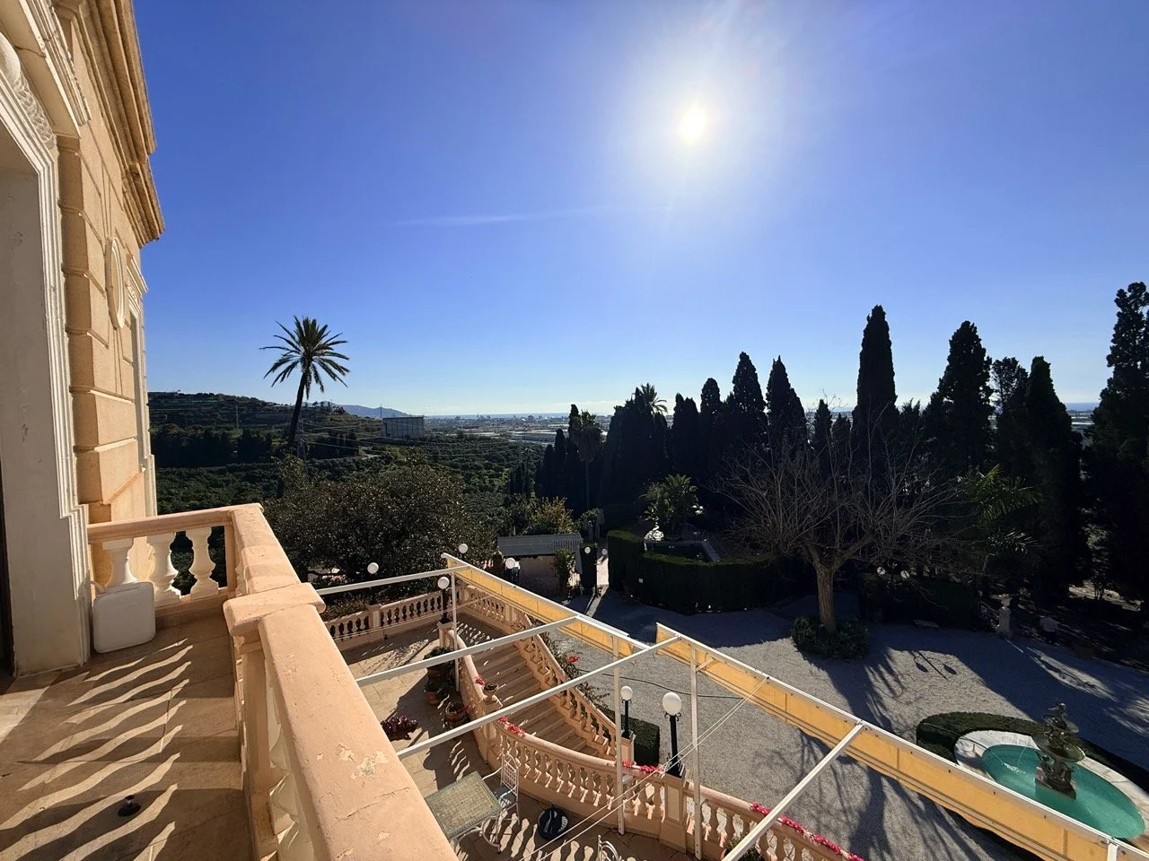 View from a balcony of a large estate or mansion, showing a garden with a fountain, trees, and an expansive landscape under a clear blue sky with the sun shining brightly.