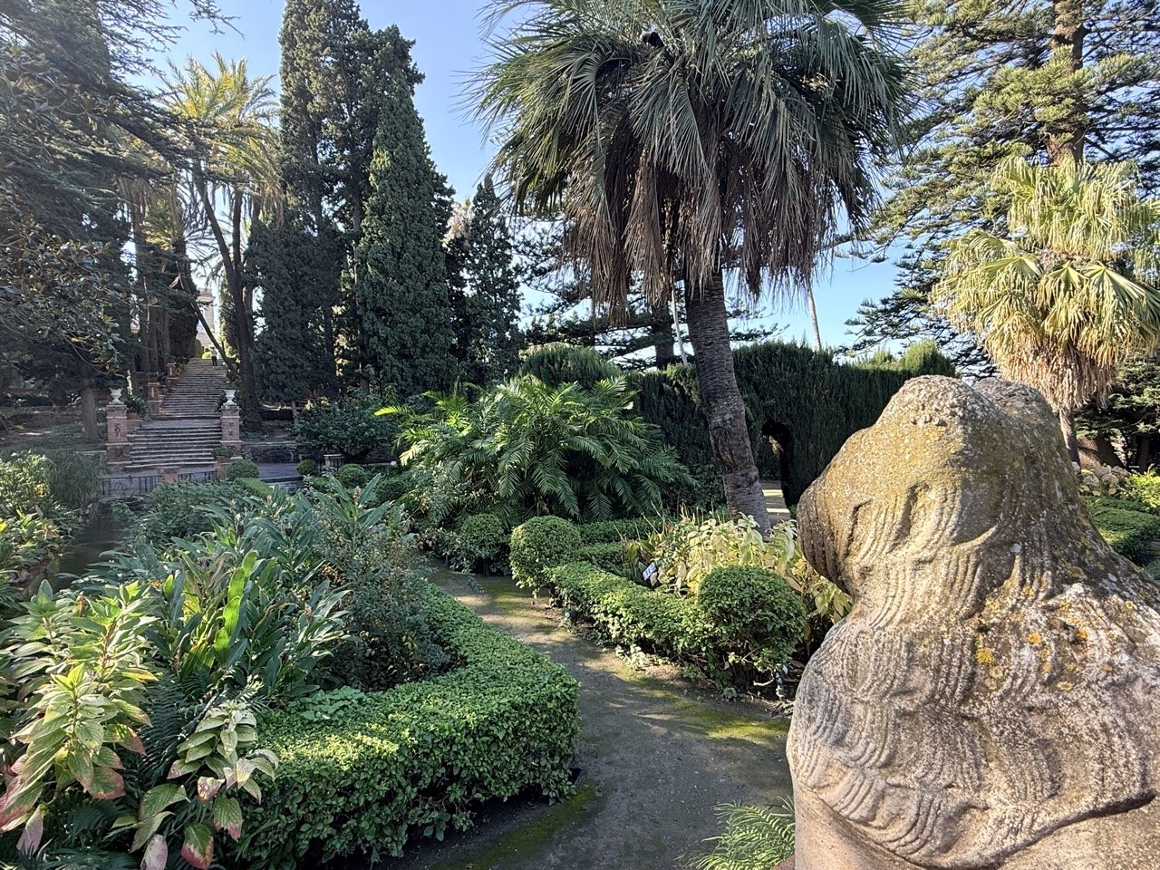 A lush garden with various trees, shrubs, and plants, including palm trees, with a stone lion sculpture in the foreground and a staircase in the background.