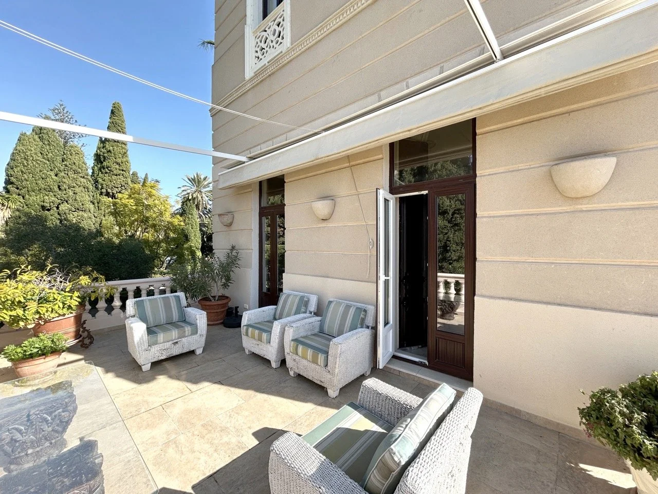 Outdoor balcony with white wicker chairs with green striped cushions, potted plants, and trees in the background under a clear blue sky.