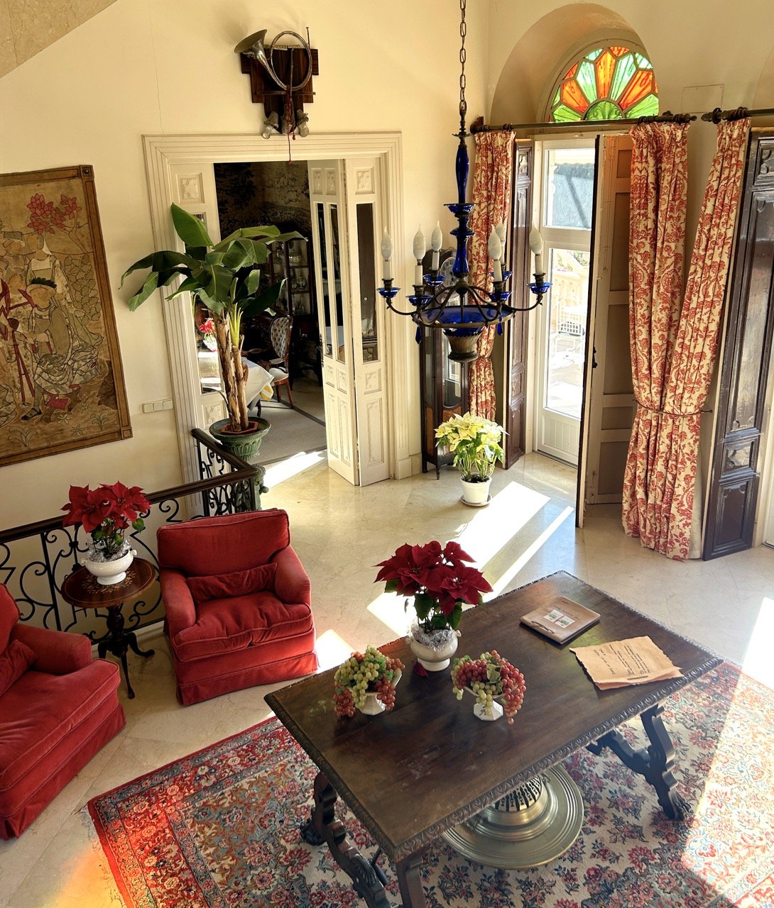 Elegant living room with red armchairs, a wooden table with floral arrangements, chandelier, large potted plant, colorful stained glass window, and decorative curtains.