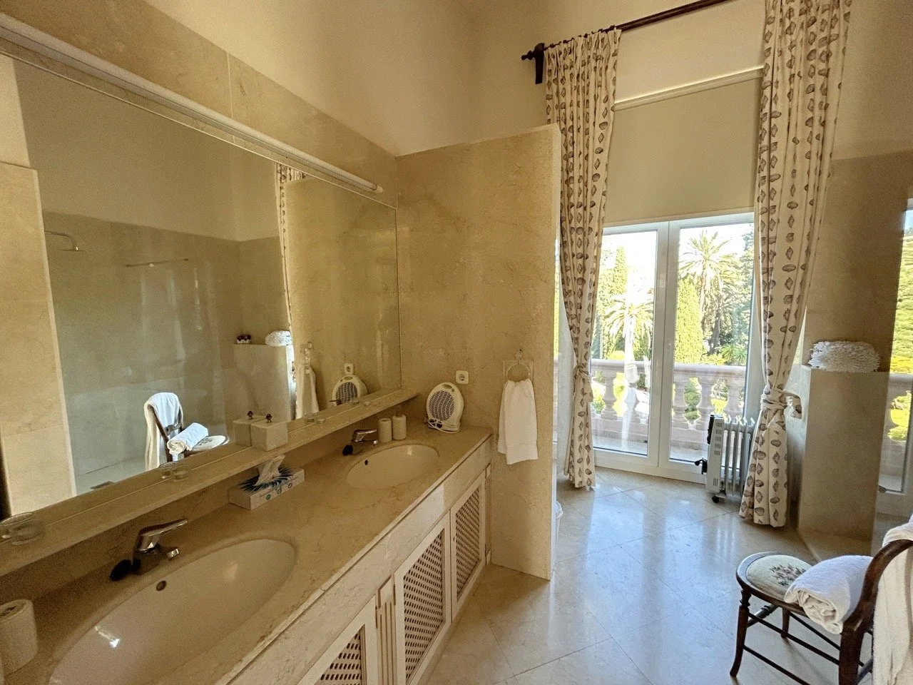 Luxurious bathroom with a double sink vanity, large mirror, and a view of a balcony and outdoor greenery through glass doors.