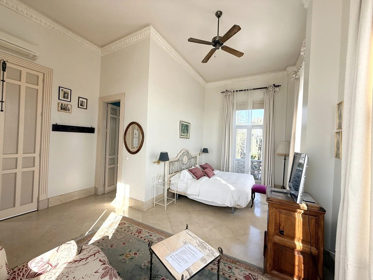Bright hotel room with white walls, a bed with white bedding and pink pillows, a wooden headboard, a small TV on a wooden stand, a window with white curtains, a ceiling fan, and a patterned area rug.