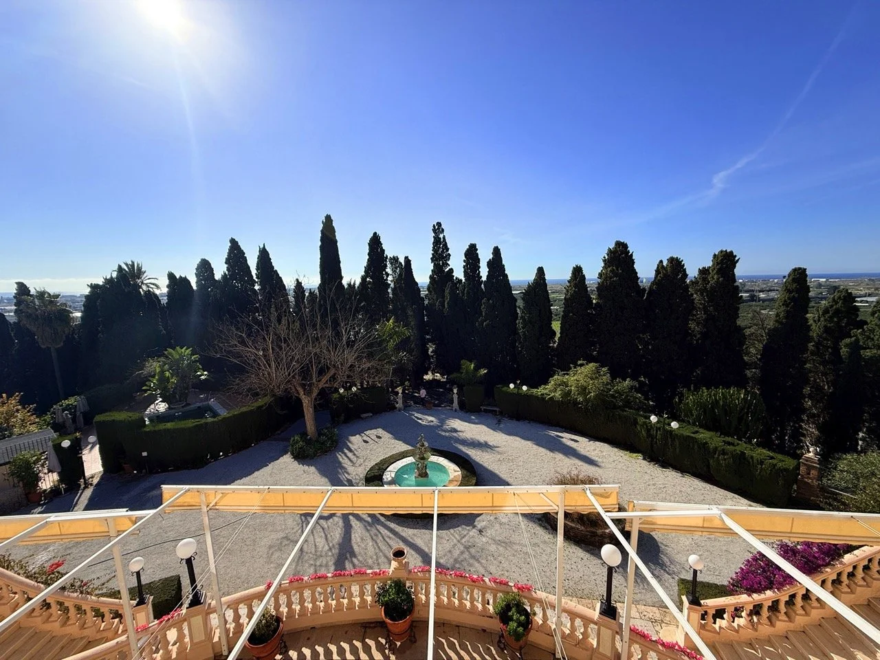 A sunny outdoor scene viewed from a building balcony with potted plants and a curved staircase. In the background, there is a garden with a fountain, tall trees, and an open landscape under a clear blue sky.