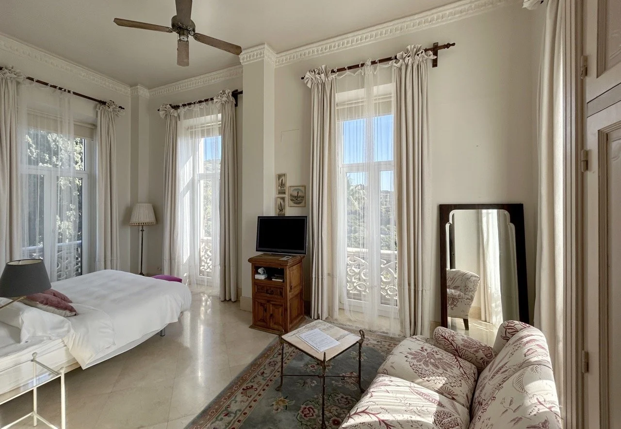 Bright bedroom with large windows, cream curtains, bed, small side table, TV on wooden stand, floral armchair, standing mirror, and ceiling fan.