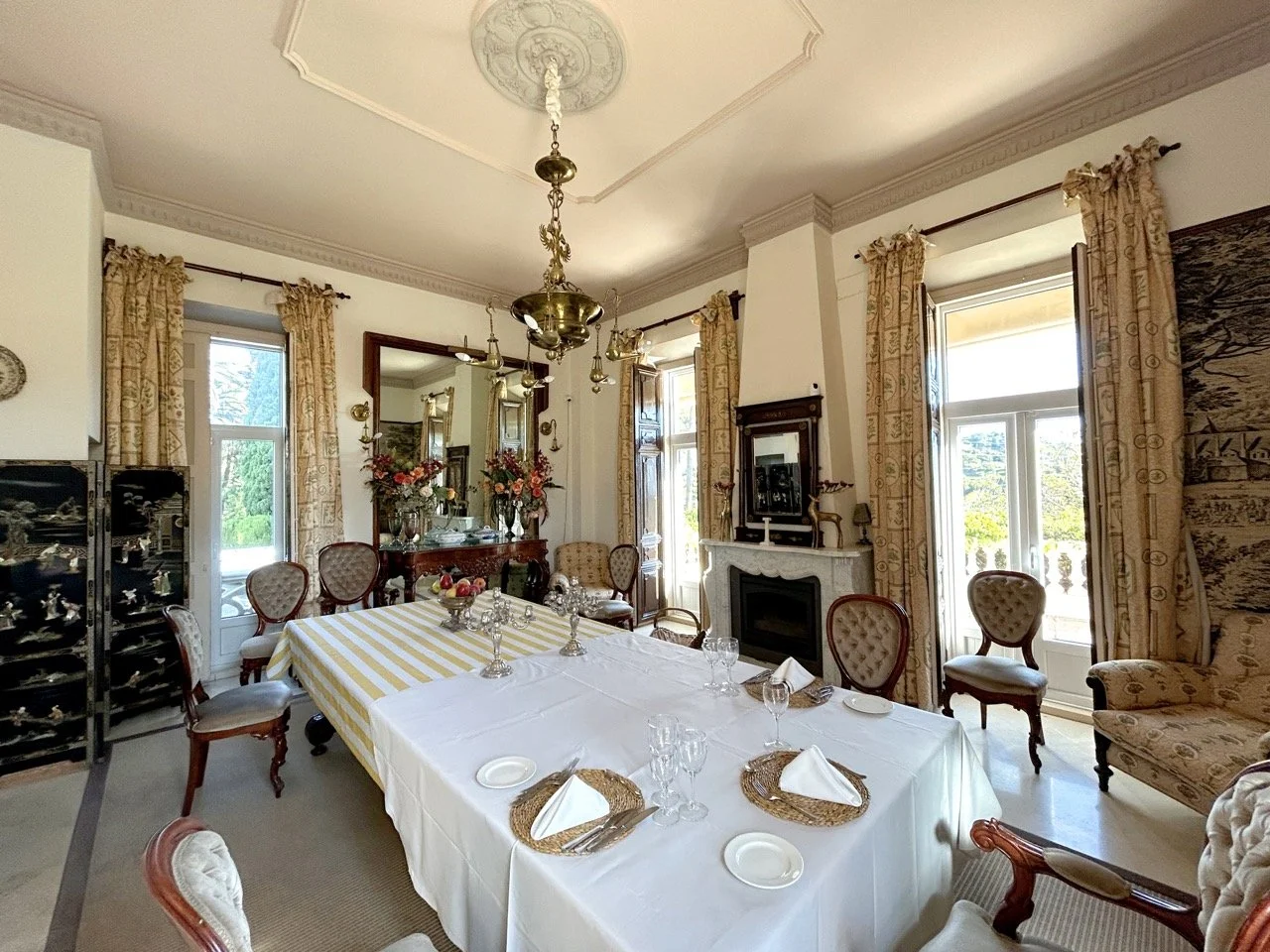 Elegant dining room with sunlight through large windows, a long table with a white tablecloth, set with plates, glasses, and napkins, surrounded by vintage chairs, floral arrangements, and ornate curtains.