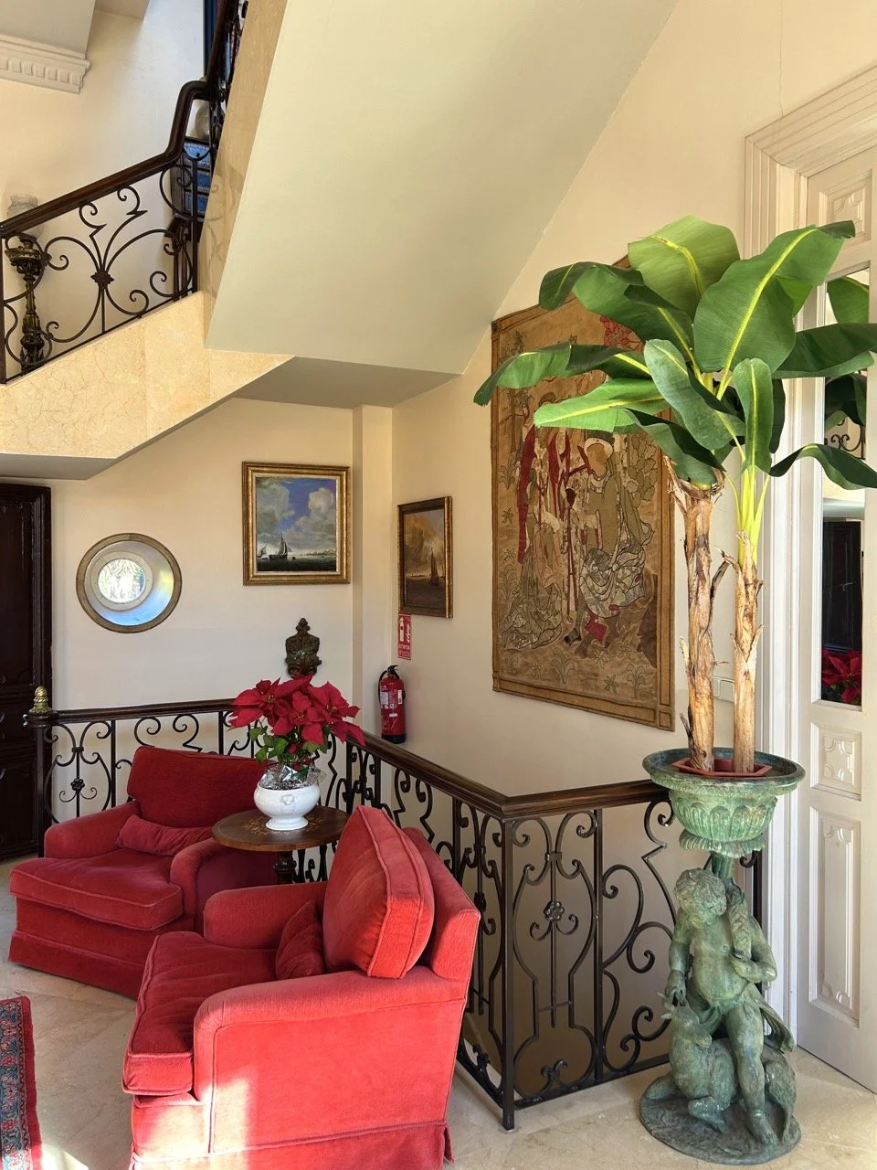 Interior of a house featuring red armchairs, a small wooden table with a poinsettia plant, framed paintings on cream-colored walls, a decorative tapestry, and a large potted plant on an ornate sculpture base near a staircase with a wrought iron raili