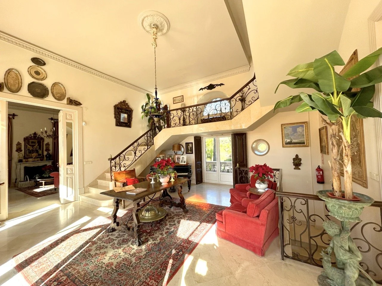 Bright living room with a staircase, a large indoor plant, red chairs with poinsettia plants, a wooden table, and framed artwork on the walls.