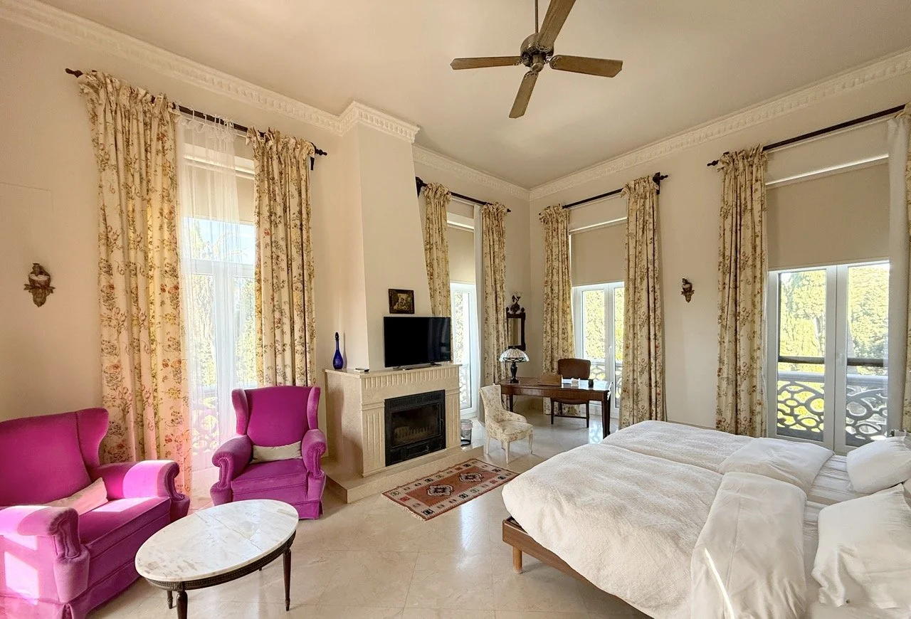 Bright hotel room with two beds, pink armchairs, a fireplace with a TV, a desk with a chair, and large windows with floral curtains overlooking a balcony.