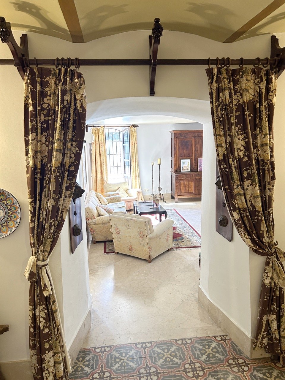 View through a doorway with floral curtains into a living room with a sofa, armchair, rug, wooden furniture, and large window with curtains.