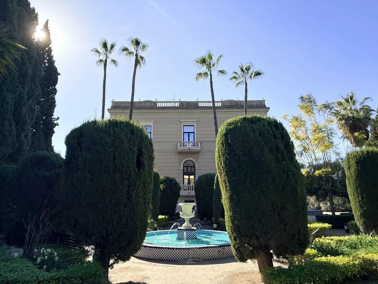 A grand estate with a symmetrical garden featuring tall palm trees, a decorative round fountain, and manicured bushes in front of a large building under a clear blue sky.
