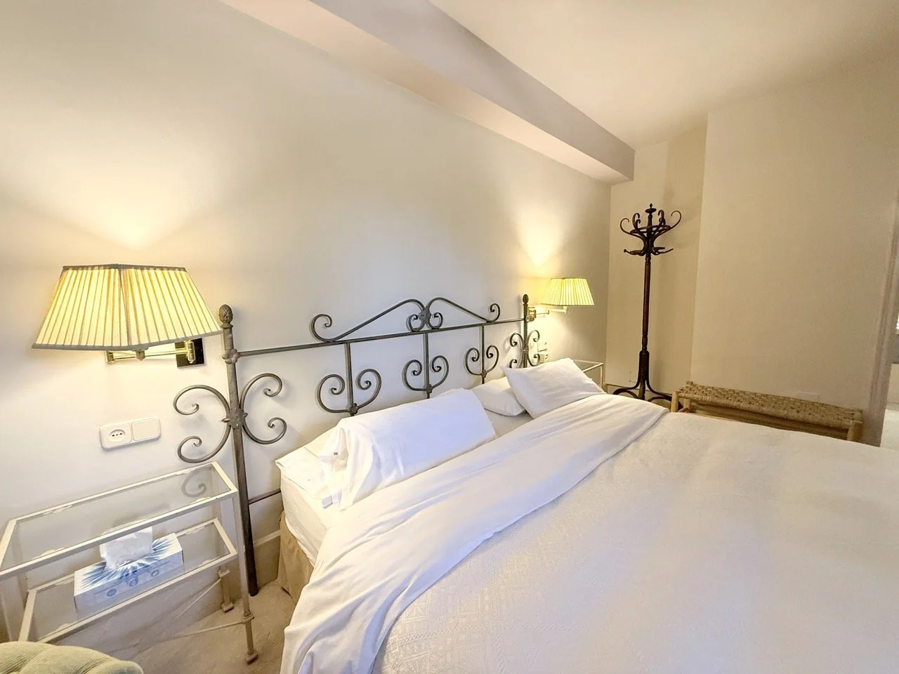 A cozy hotel bedroom with a metal bed frame, white bedding, bedside lamps, a coat rack, and a wicker bench.