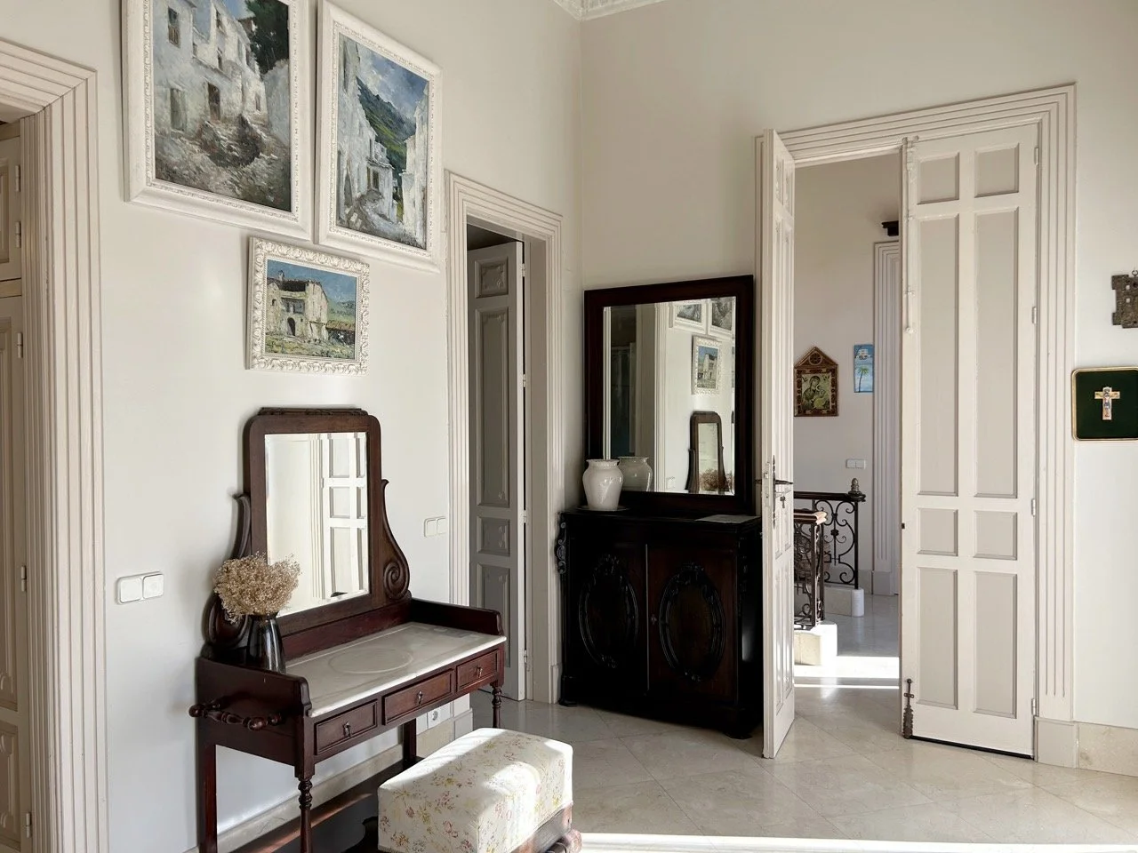 A corner of a room with white walls decorated with framed landscape paintings, a vintage wooden mirror with a vase of dried flowers, a dark wooden sideboard with two white vases, and an open double door leading to a staircase with decorative railing.