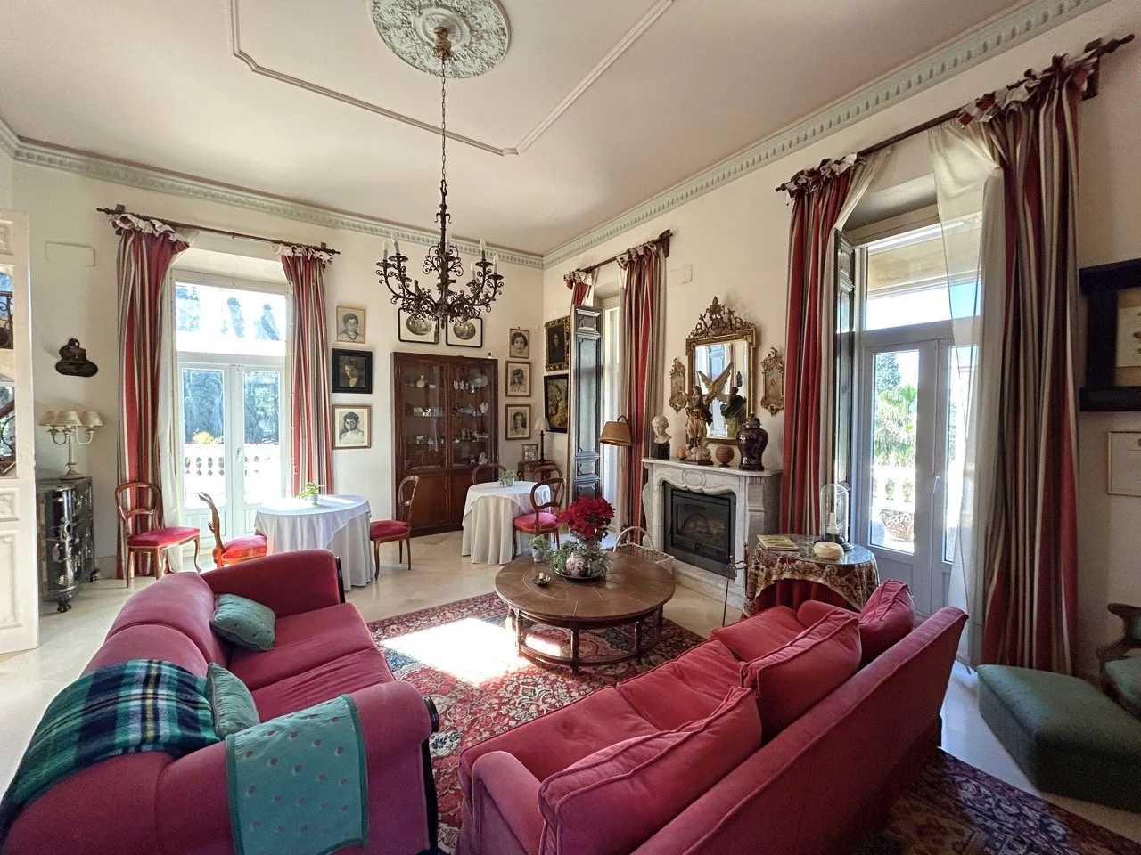 Elegant living room with pink sofas, a round wooden coffee table, a chandelier, a fireplace with gold accents, and large windows with pink and cream curtains.