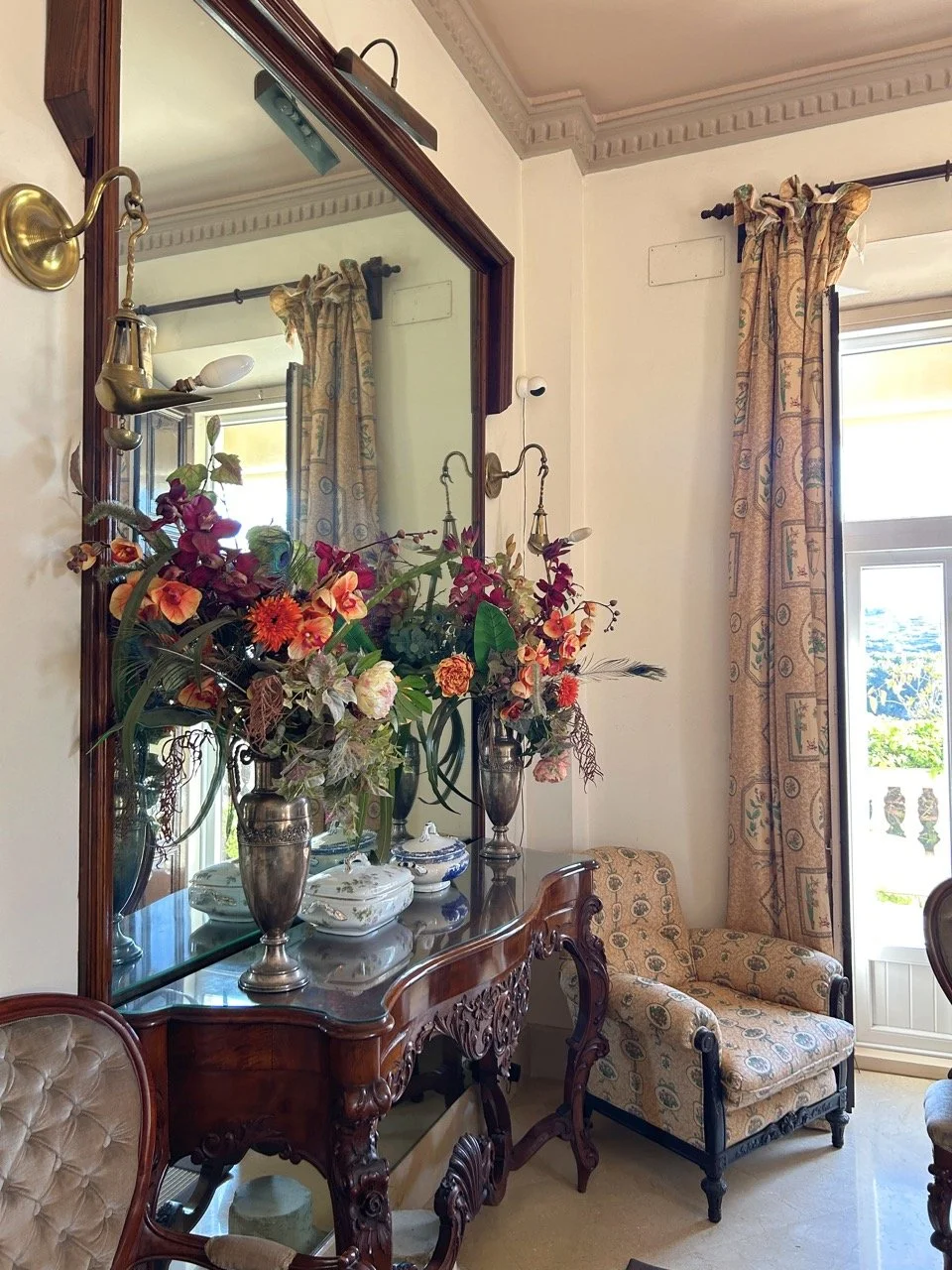 A decorated corner of a room with a wooden console table, a large mirror, a bouquet of colorful flowers in silver vases, a vintage upholstered armchair, and full-length curtains in front of a window.