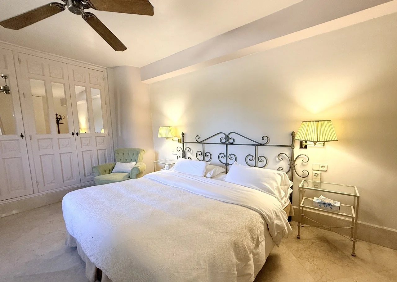 A cozy bedroom with a large bed featuring a decorative metal headboard, white bedding, two bedside lamps with green shades, a light green armchair with a white pillow, a ceiling fan, and built-in white wardrobes with mirrored doors.