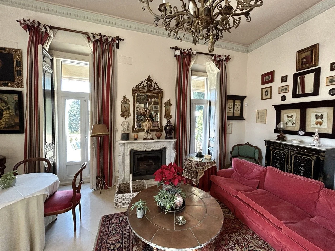 A decorated living room with a red velvet sofa, a round wooden coffee table with a poinsettia plant, a fireplace with decorative items, and multiple framed artworks on the walls. There's a chandelier above and two tall windows with curtains.
