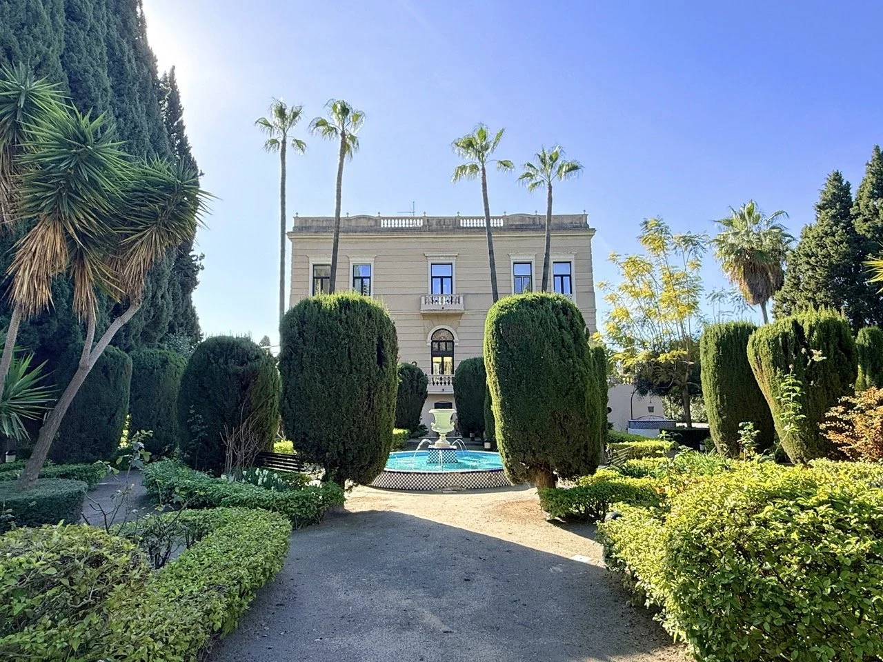 A landscaped garden with tall palm trees, trimmed bushes, a fountain, and a grand building in the background under a clear blue sky.