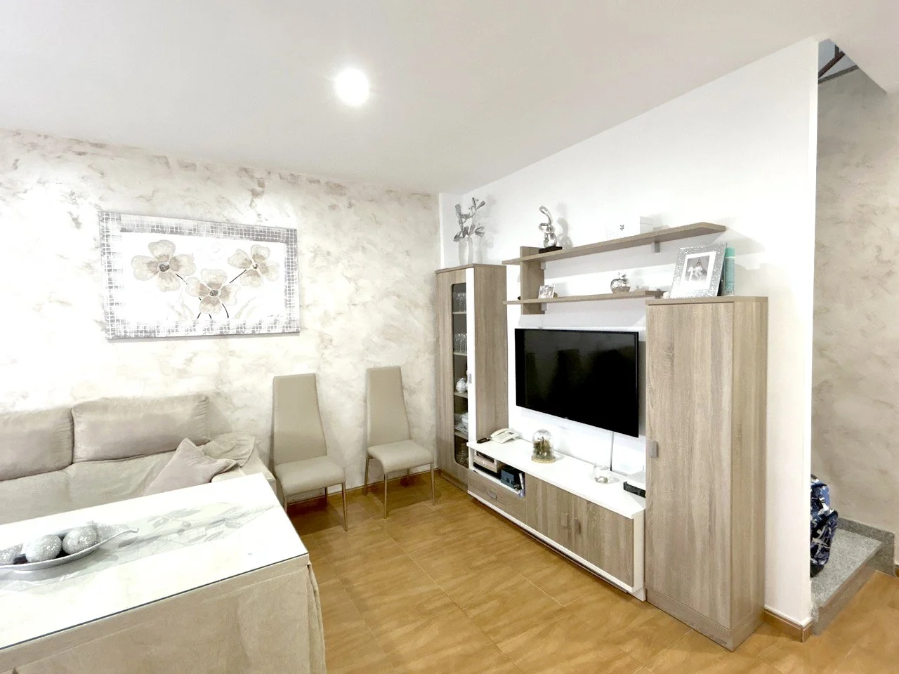 Living room with beige sofa, two beige chairs, a wall-mounted TV, wooden shelves, white textured wall, and decorative items.