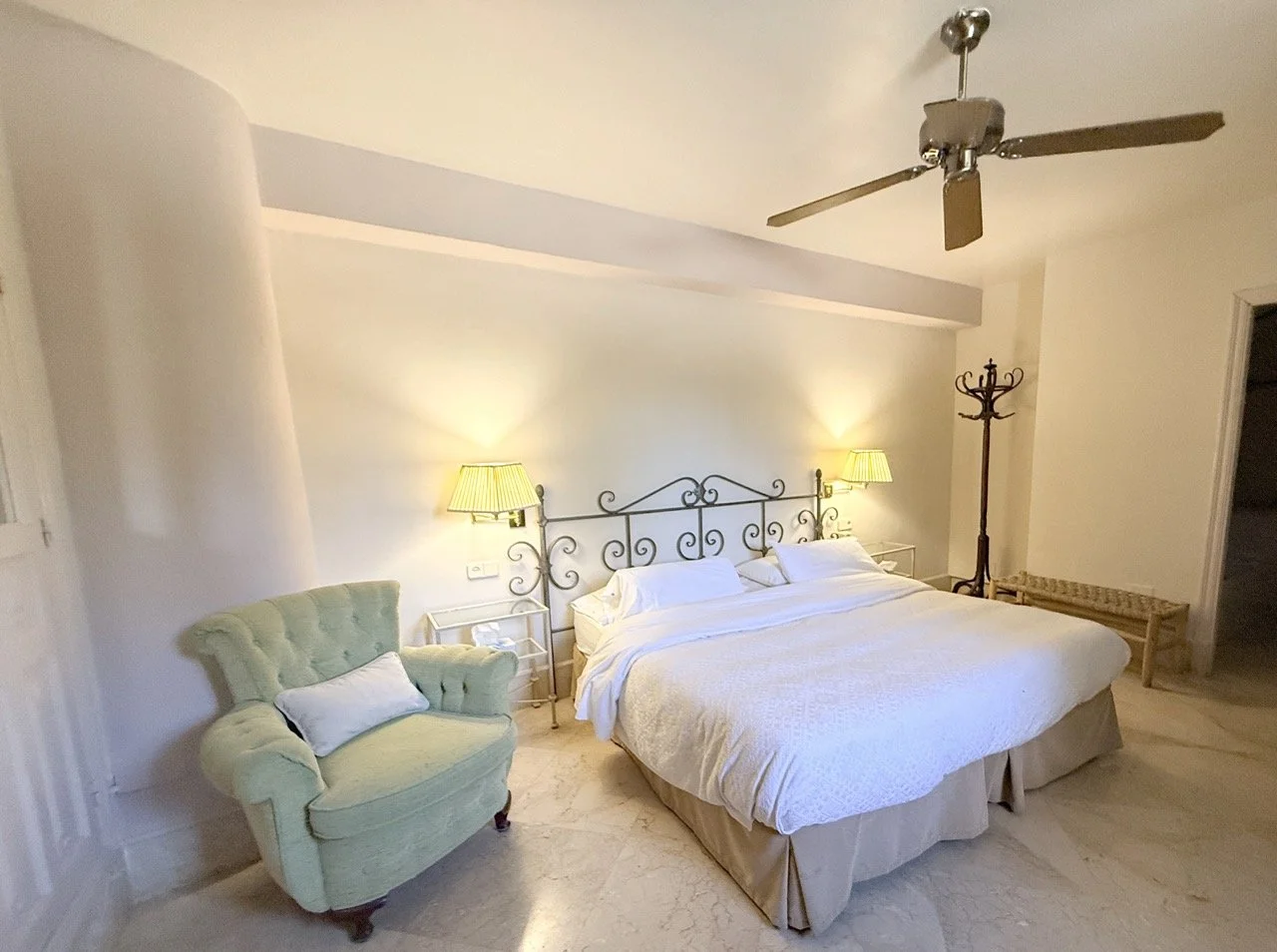 A bedroom with a king-sized bed, two bedside tables with yellow lamps, a vintage green armchair with a pillow, a coat rack, and a ceiling fan.