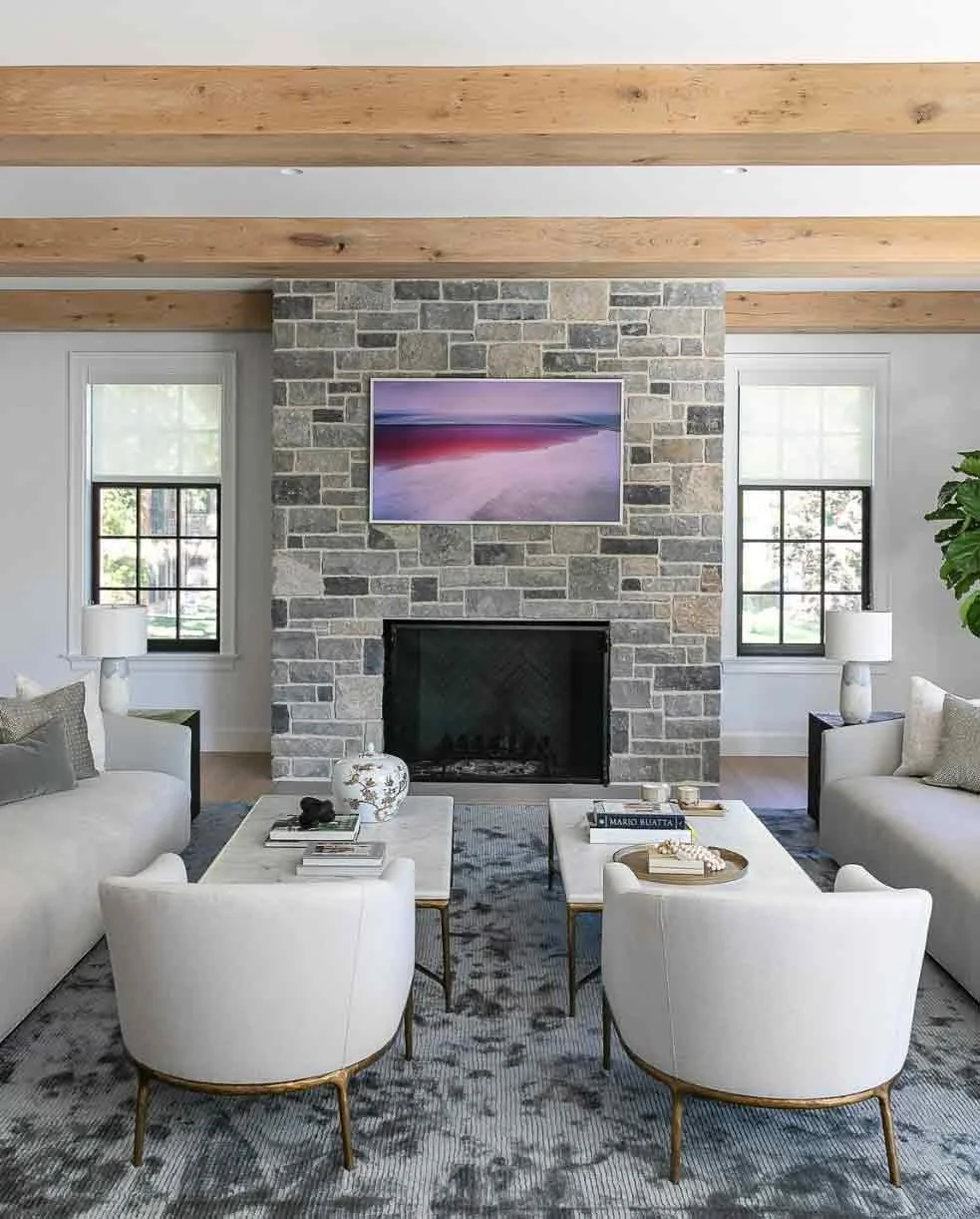 Living room with a stone fireplace, wall-mounted TV, two windows with blinds, beige sofas, round armchairs, side tables, table lamps, and decorative items on the coffee tables.