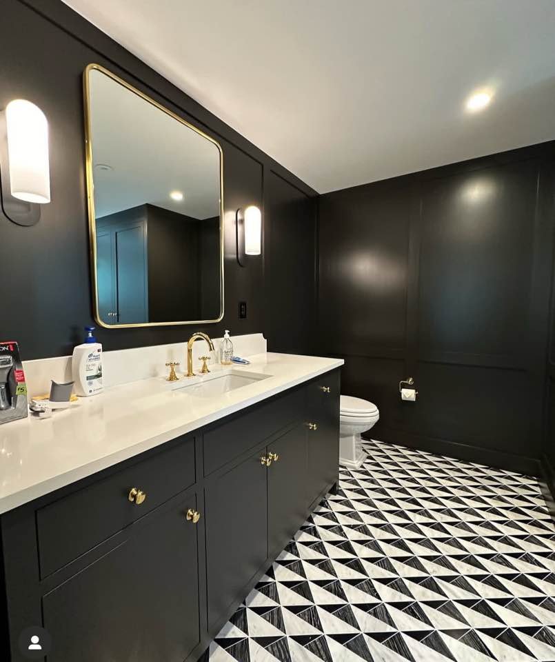 Modern bathroom with black walls and a black vanity with gold hardware. White countertop with a sink, gold faucet, and various toiletries. Large mirror with gold trim, flanked by two vertical wall sconces. Black-and-white patterned floor tiles. Toilet in the corner, framed by the black wall.