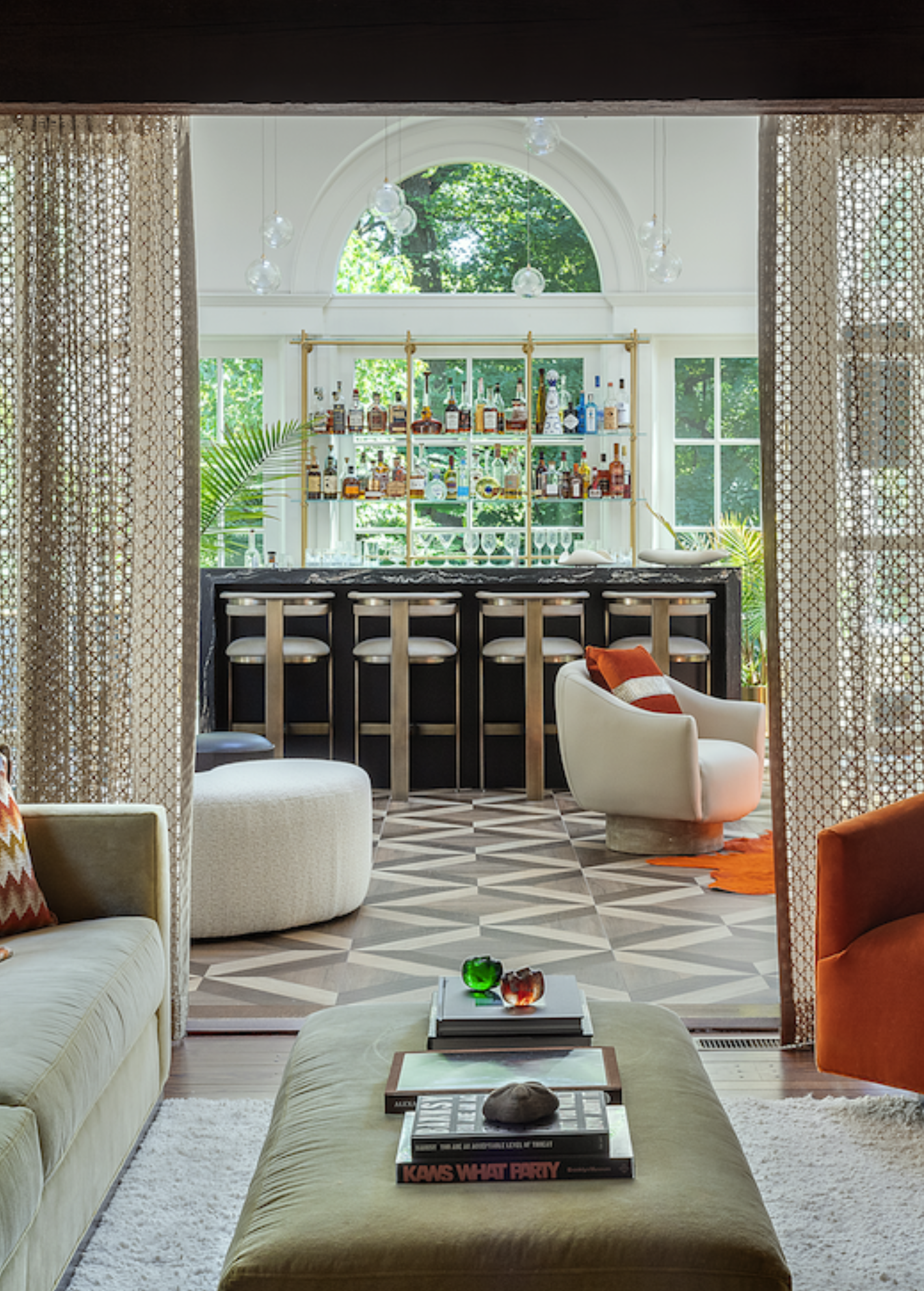 Living room with a view of a home bar, featuring a black counter and bar stools, surrounded by large windows and lush greenery, decorated with patterned curtains, colorful pillows, and books on a coffee table.