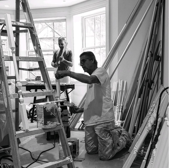 Two men working on a woodworking project in a well-lit room with large windows. One man is standing at a table in the background, while the other is kneeling and painting or staining a wooden spindle.