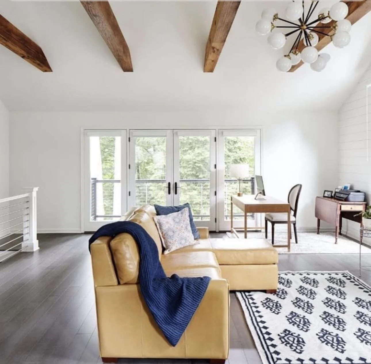 Cervone Living room with yellow sectional sofa, navy blue throw blanket, pillows, wooden ceiling beams, modern chandelier, large windows with balcony, white walls, dark wood flooring, desk with computer, side table with printer, patterned rug.