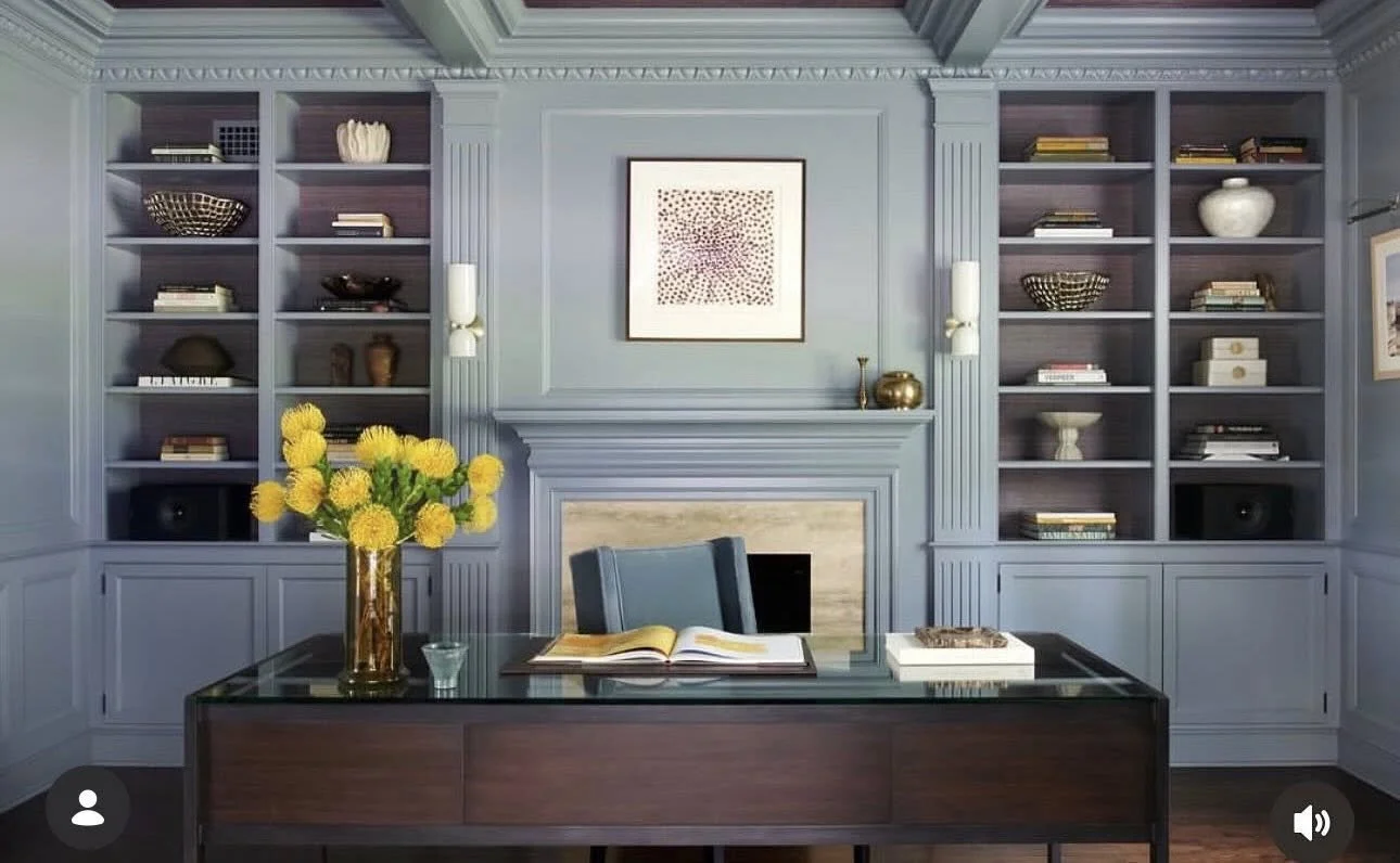 Cervone Painting Services Philadelphia Main Line - Office with blue built-in bookshelves, a central framed artwork, and a wooden desk with a vase of yellow flowers, an open book, and a tray.