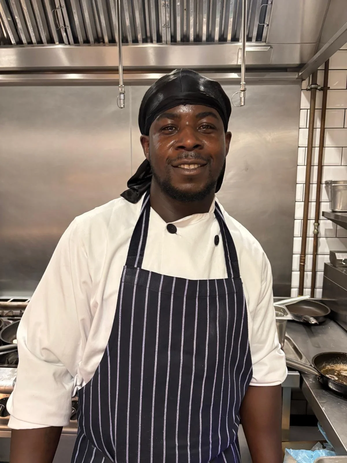 A smiling chef wearing a black headscarf, white chef's coat, and a dark striped apron standing in a professional kitchen.