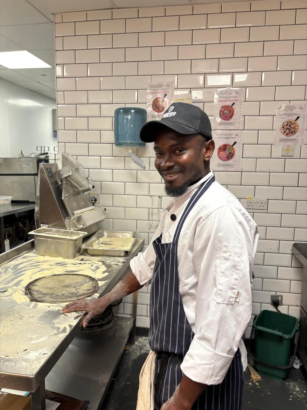 A chef in a striped apron and black cap working at a pasta station with flour and dough in a commercial kitchen.