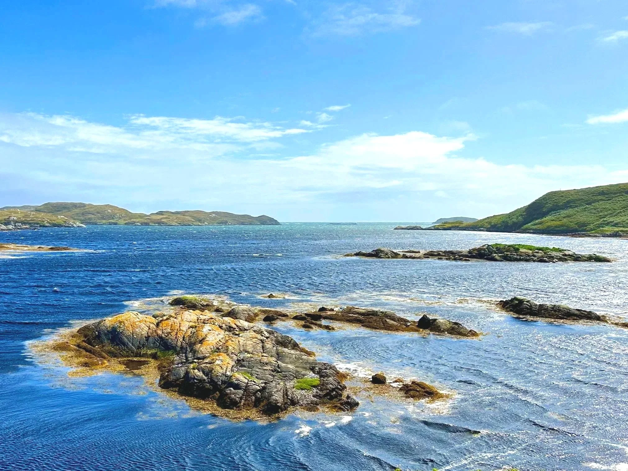 Coastal scene with blue water, rocks, and green hills under a partly cloudy sky.