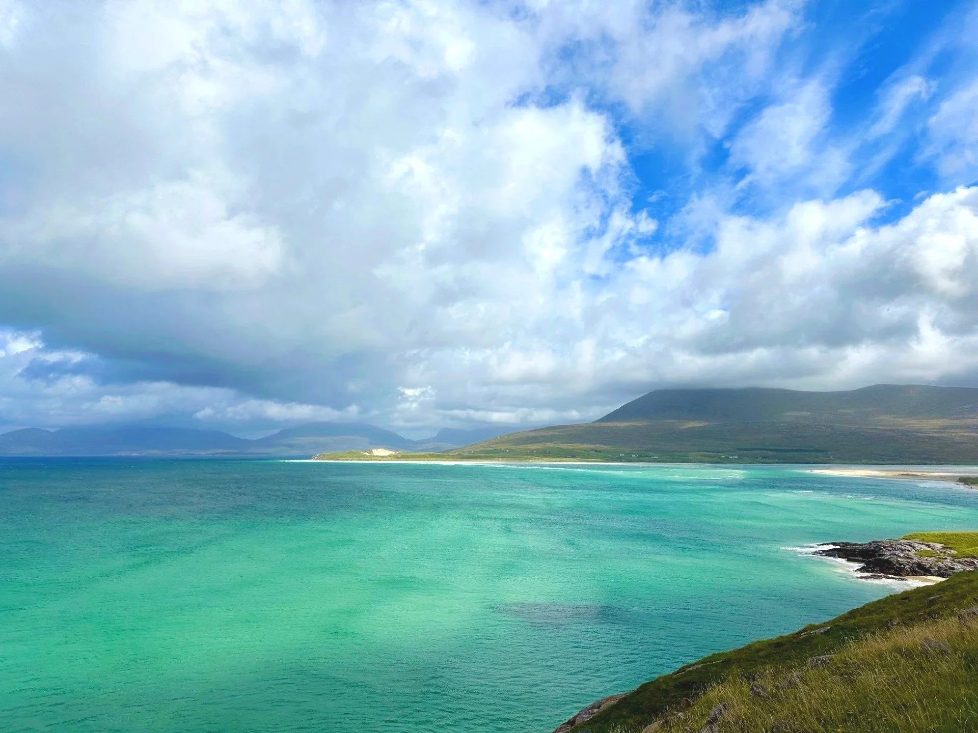 A coastal landscape with turquoise waters, green grassy hills, and mountains in the background under a partly cloudy sky.