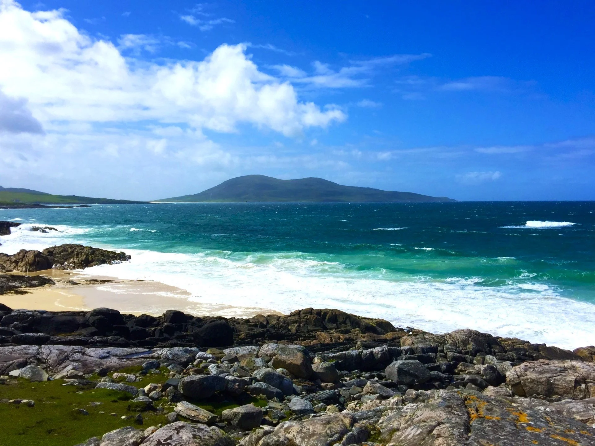 Scenic view of a rocky coastline with green grass in the foreground, ocean waves crashing on the shore, and a mountain in the distance under a partly cloudy blue sky.
