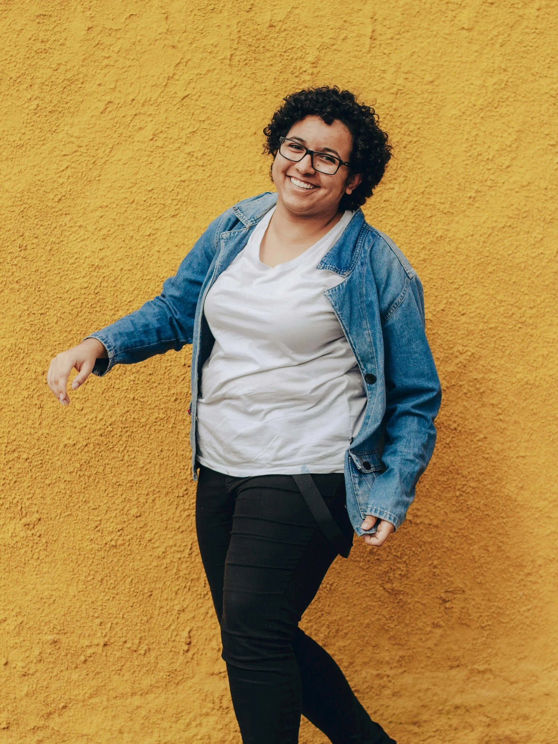 Person with curly hair and glasses smiling, wearing a white t-shirt and denim jacket, standing against a yellow textured wall.