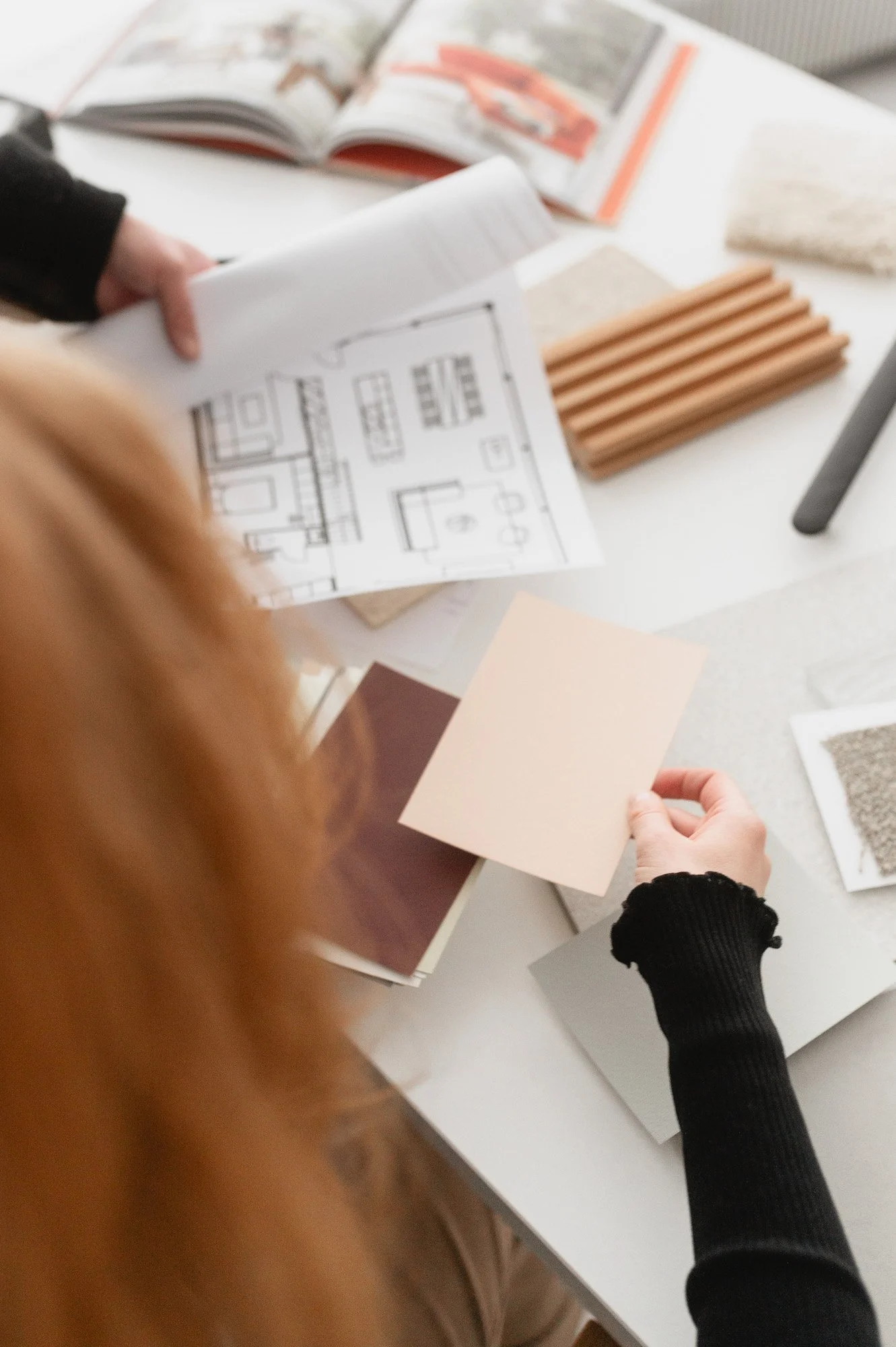 Kuva ihmisistä, jotka tutkivat sisustussuunnitelmia ja näytteitä, pöydällä on rakennuspiirustuksia, väritarroja ja eri materiaalinäytteitä.