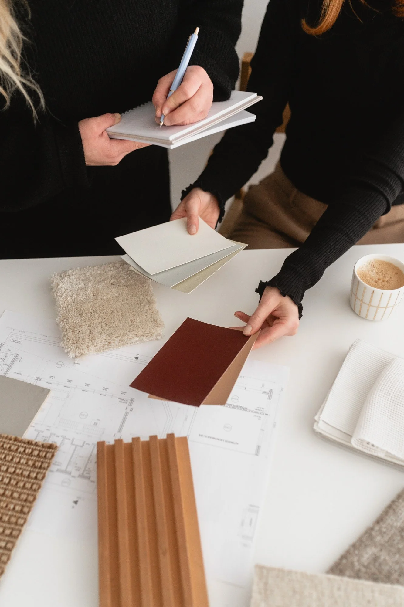 Kaksi ihmistä käsittelee sisustusmateriaaleja kuten maaleja, kankaita ja puusepäntarvikkeita pöydän ääressä, jolta näkyy lattiasuunnitelma. Brändikuvaus, sisustussuunnittelu