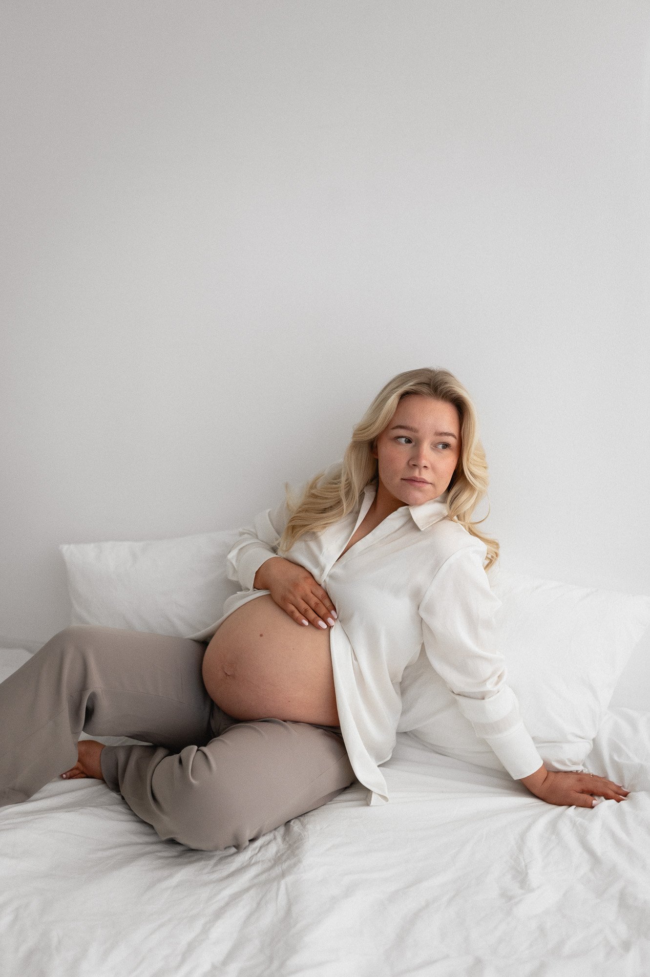 Raskaana oleva nainen istuu sängyllä, silmät katseessa poispäin, valkoisen seinän edessä.