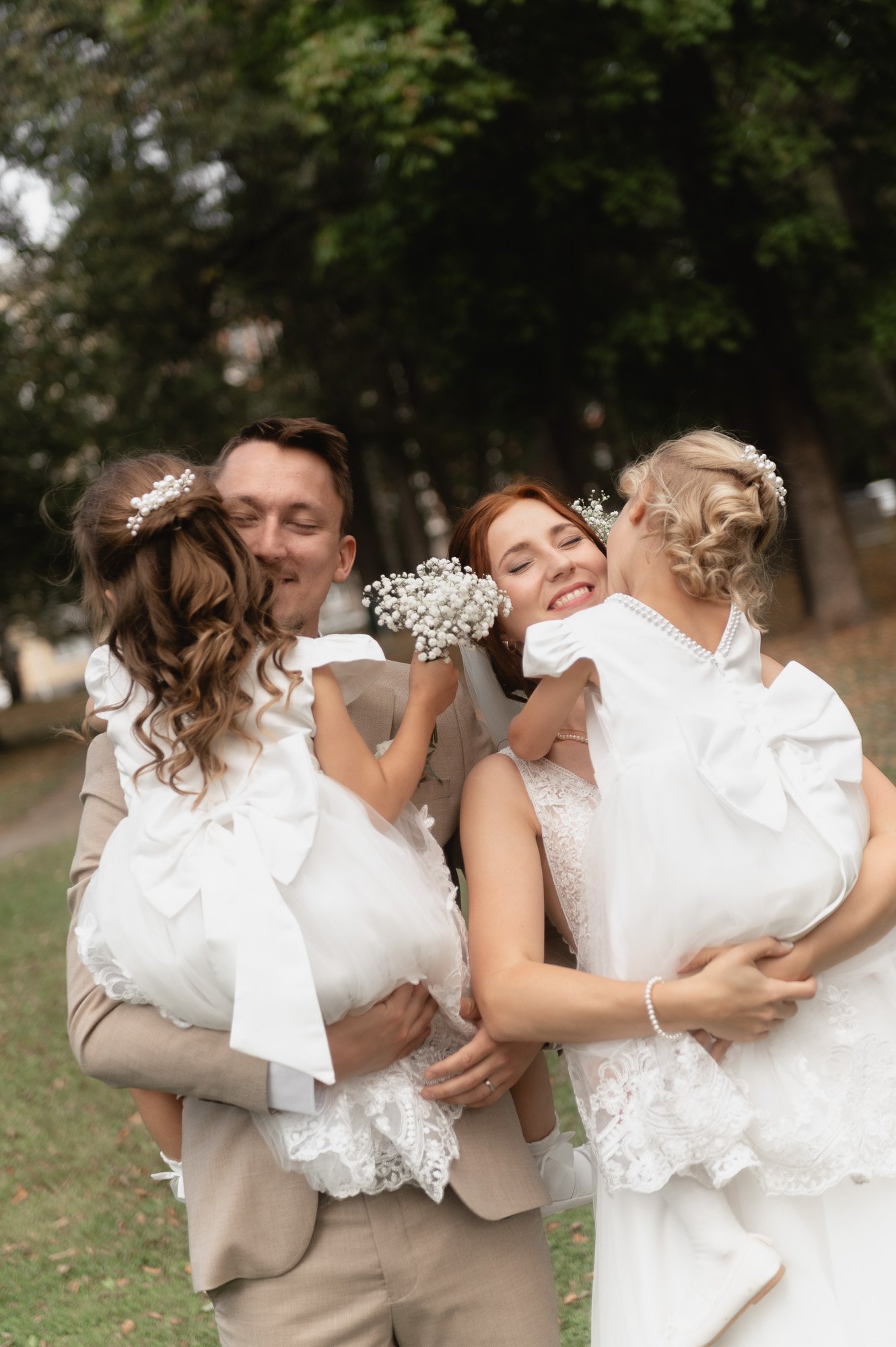 Hääpari ja heidän kaksi pientä tyttöä valkoisissa juhlamekoissa, iloisesti halaavat ja nauravat ulkona puistossa.