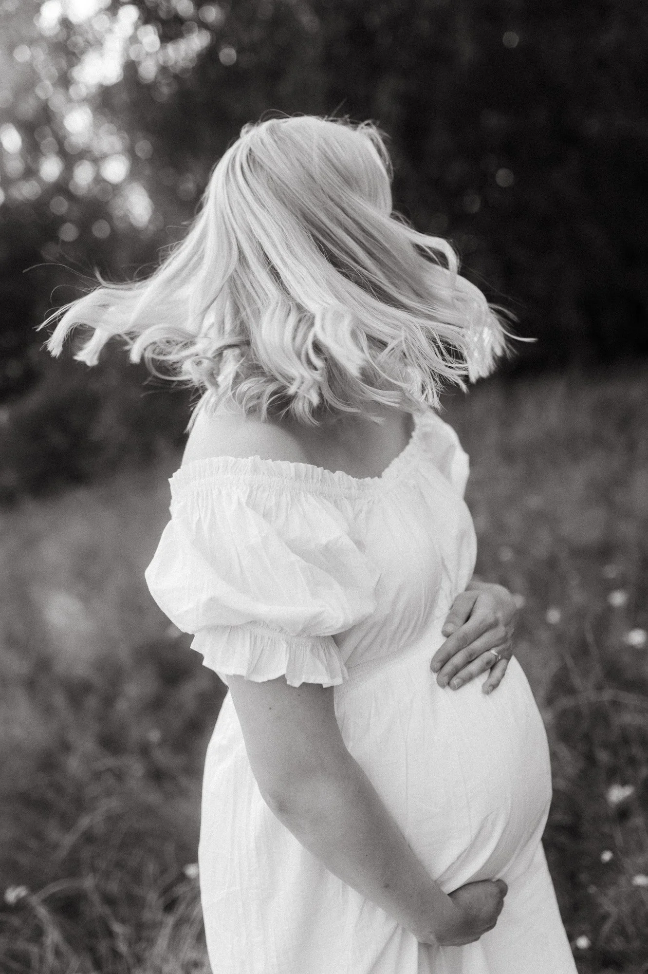 Raskaana oleva nainen vaaleassa valkoisessa mekossa, vaaleat kiharat hiukset, seisoo ulkona luonnossa, taustalla puita ja kasveja, mustavalkoinen kuva, odotusajan kuvaus