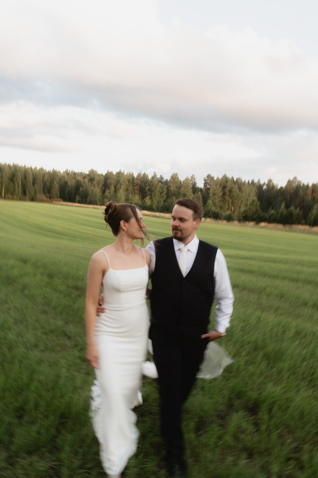 Hääpari kävelee pellolla ja katsoo toisiaan, nainen valkoisessa mekossa ja mies pikkutakissa, taustalla metsä ja pilvipoutainen taivas, kesäilta, hääkuvaus