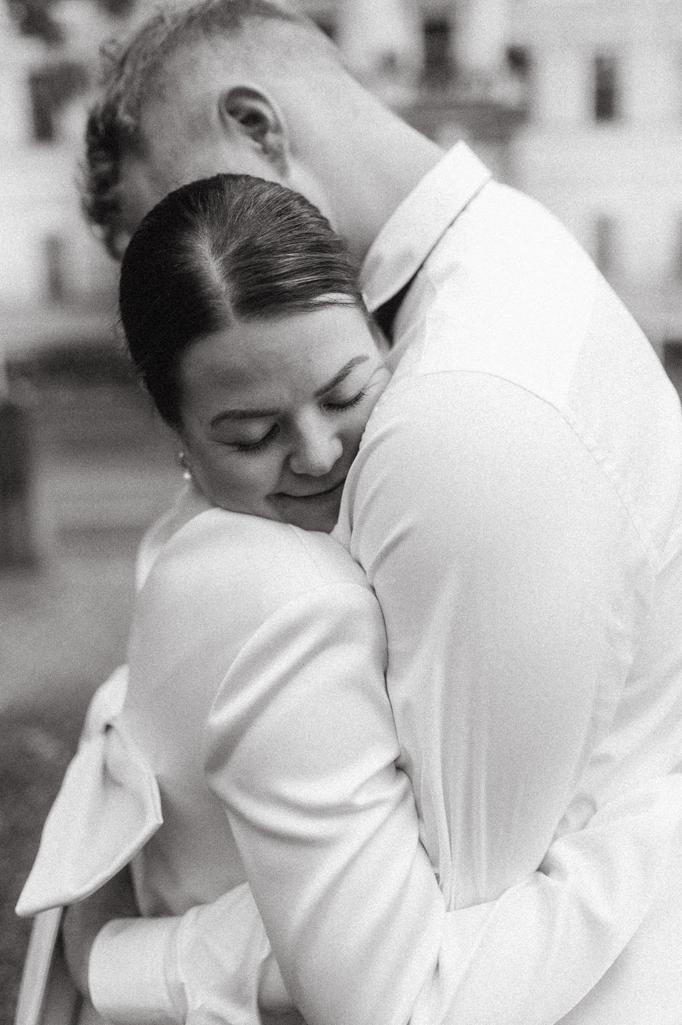 Mustavalkoinen kuva hääparista halaamassa tiukasti, hymy, taustalla vaalea rakennus, hääkuvaus