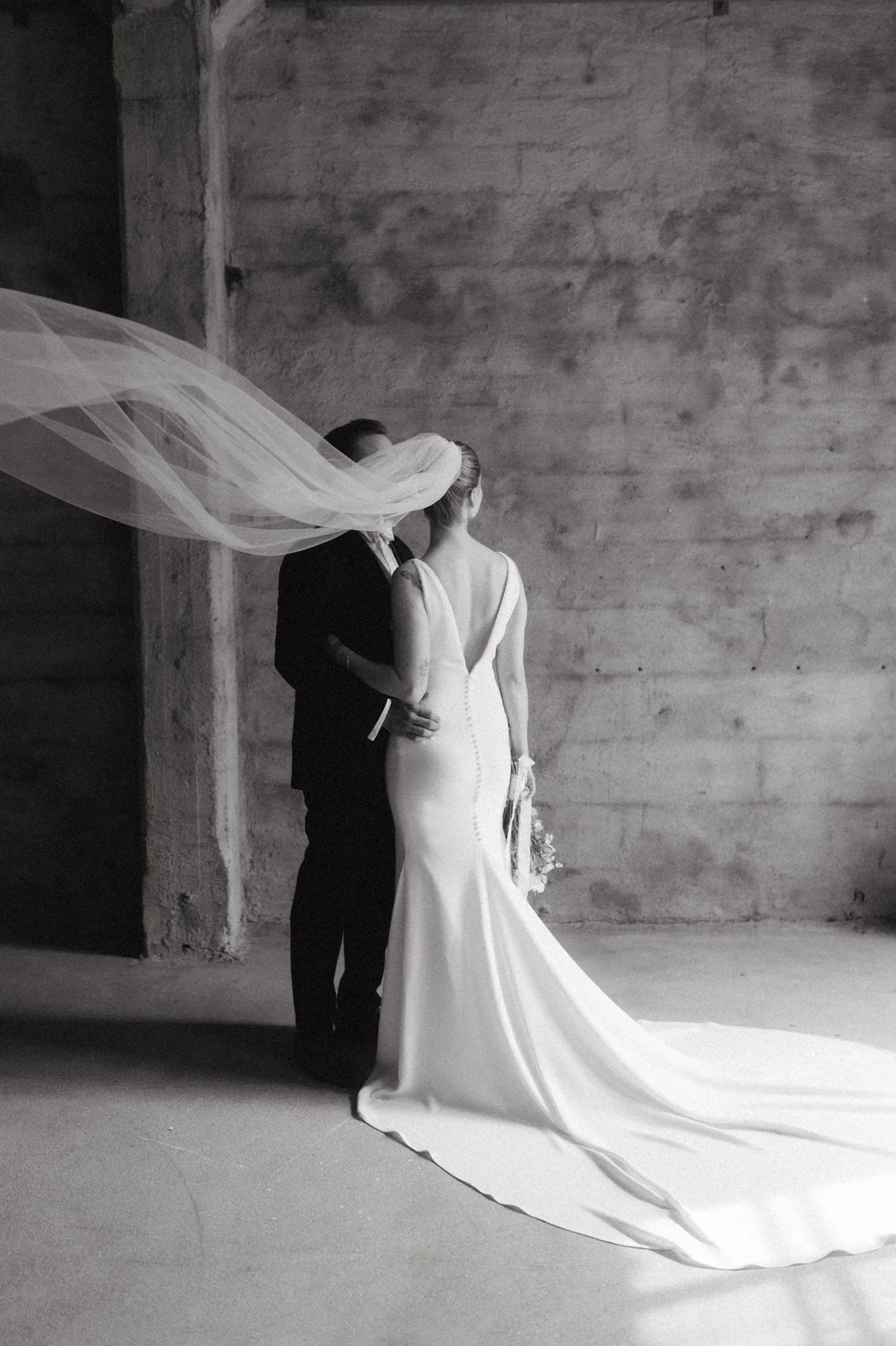 Morsiuspari, nainen häämekossa ja mies puvussa, poseeraa kahdestaan varjossa, valkoinen häämekko ja viitta, häävalo, mustavalkokuva.