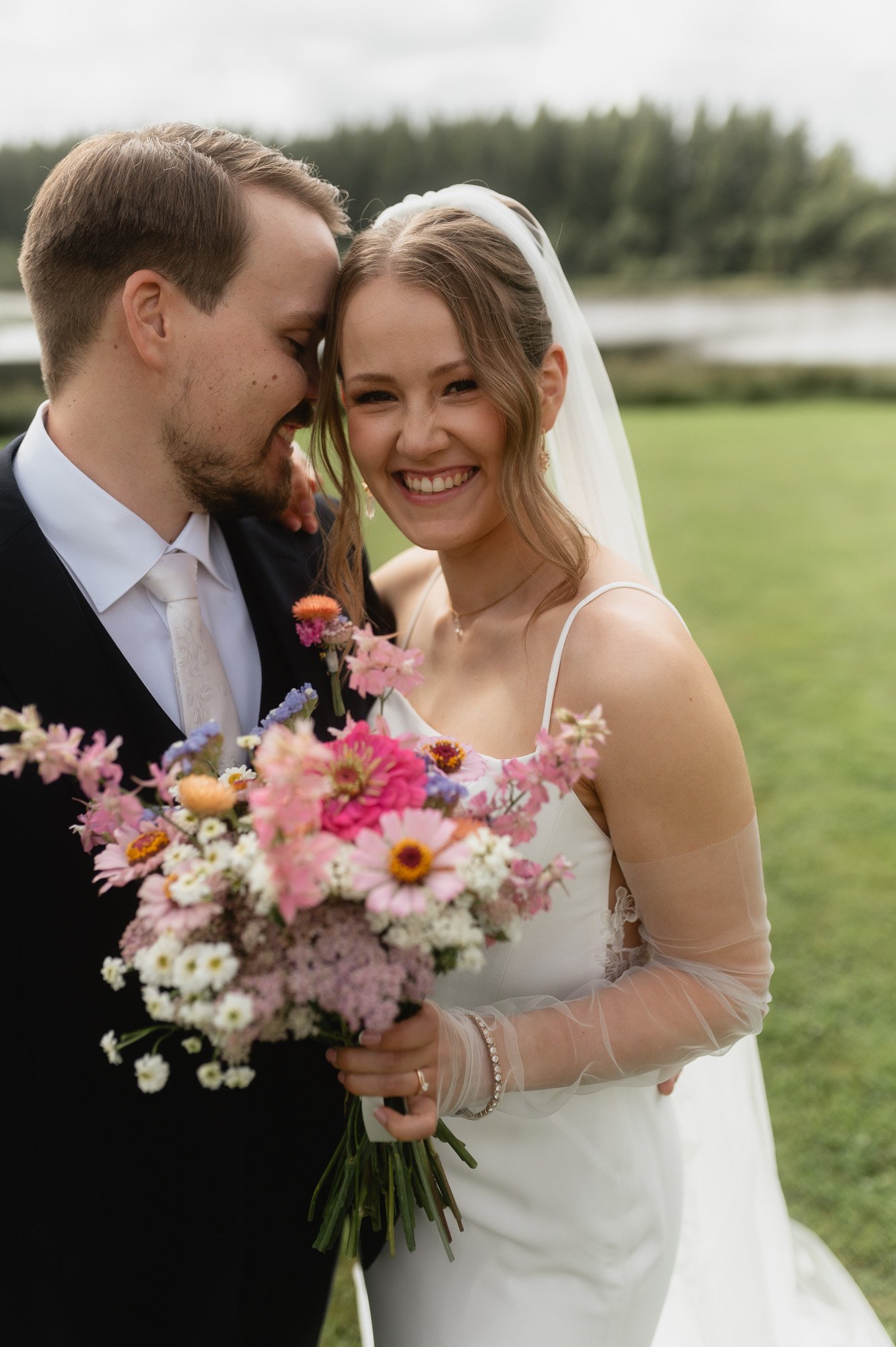 Hääpari hymyilee ja halailee toisiaan ulkona järven rannalla, morsian pitää kukkakimppua.