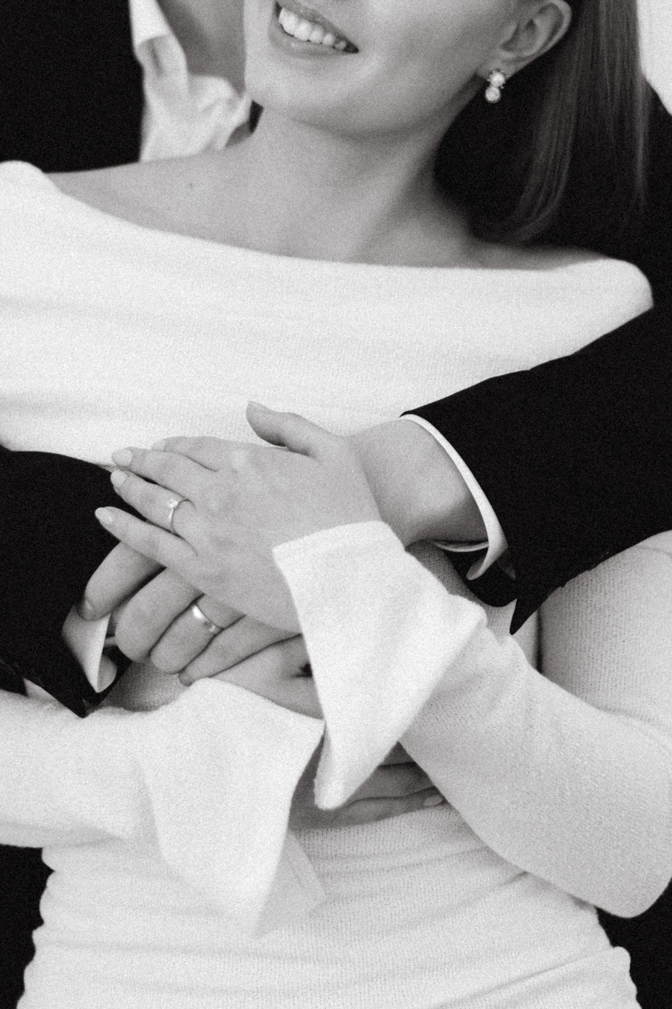 Hymyilevä nainen, jonka ylävartalo ja kädet, käyttämättömät sormukset ja korvakorut näkyvät, mustavalkoisessa kuvassa.