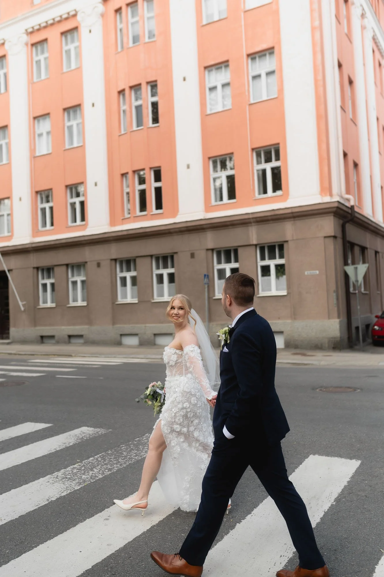 Hääpari kävelee käsi kädessä risteyksessä kaupungin kadulla, taustalla monikerroksinen, vaaleanpunainen vanha rakennus.