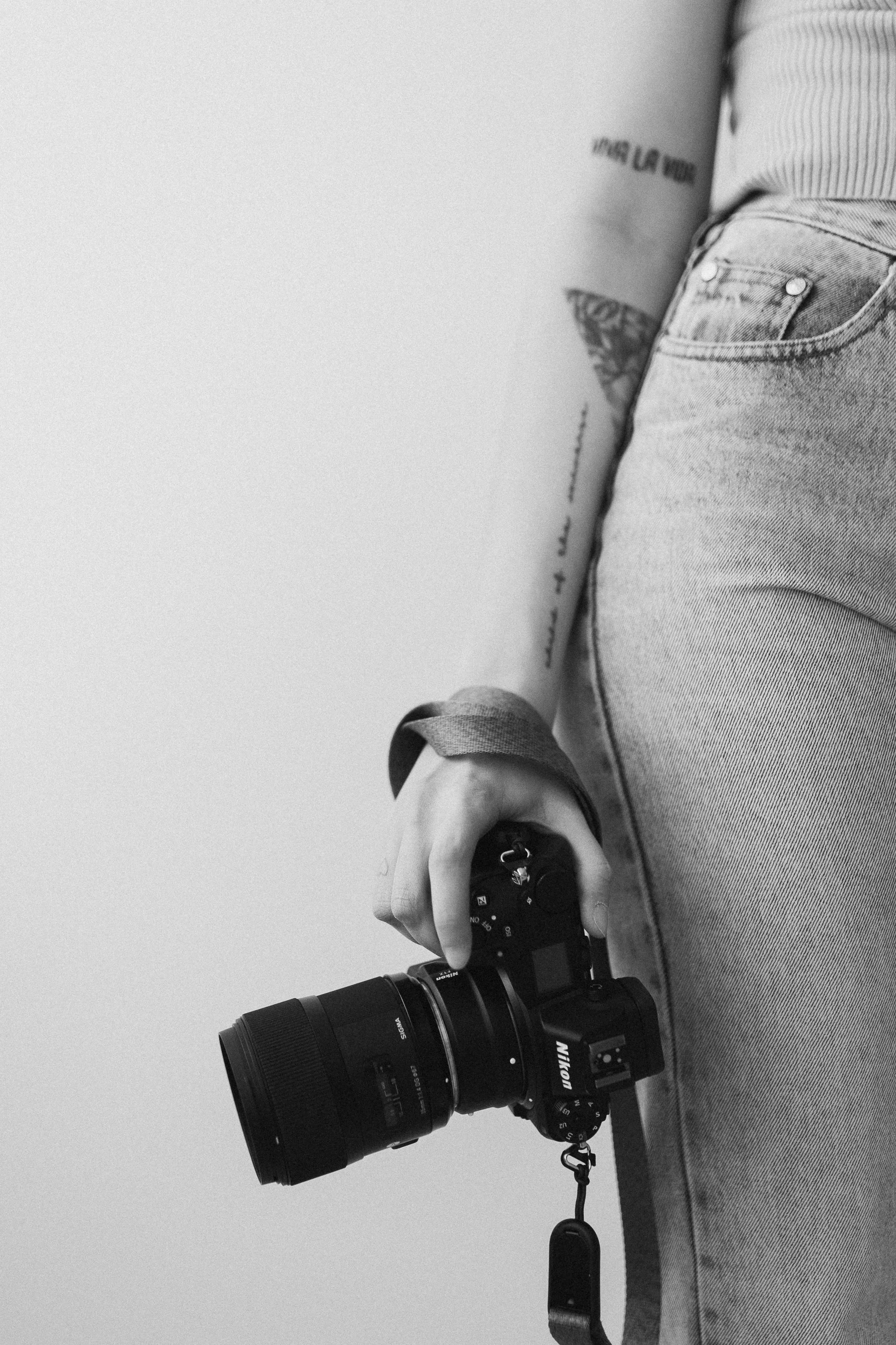 Valokuvaaja Kuopio, mustavalkoinen valokuva henkilöstä, joka pitää kameraa ja on pukeutunut farkkuihin ja puseroon.